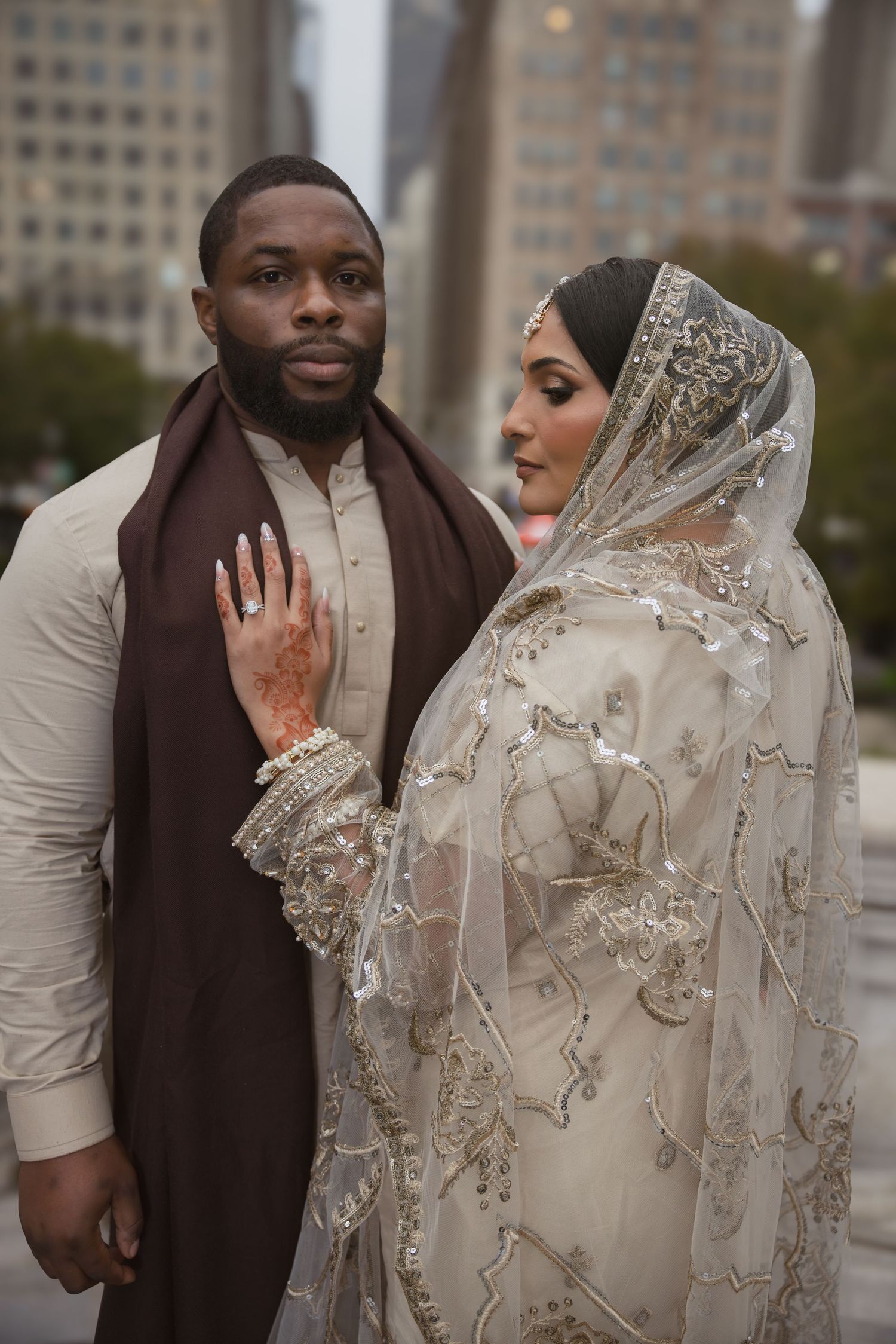 Bride in embroidered white veil and groom in cream sherwani with brown scarf pose in city.