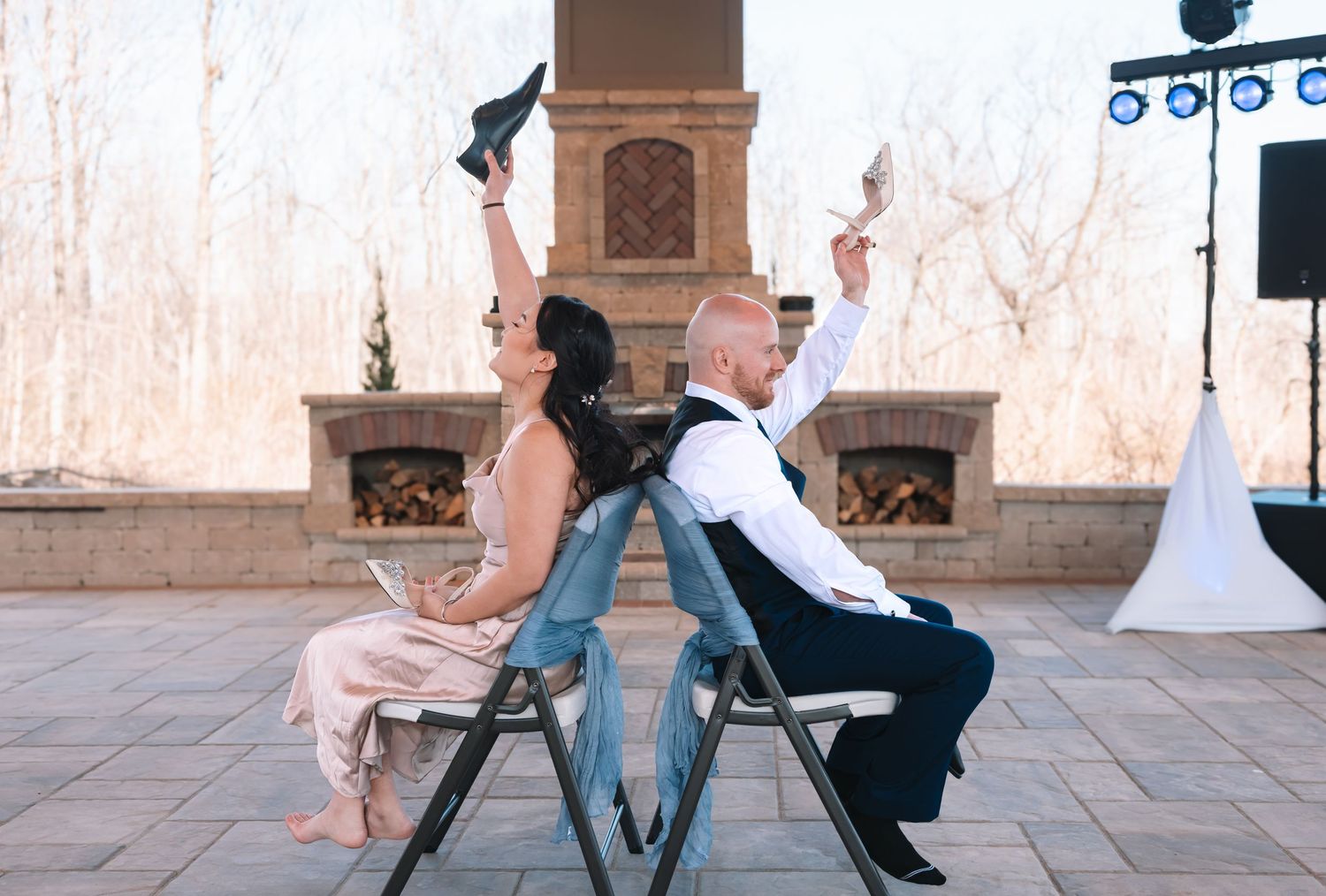 Bride and groom sit back-to-back on chairs raising shoes playfully at outdoor wedding reception.