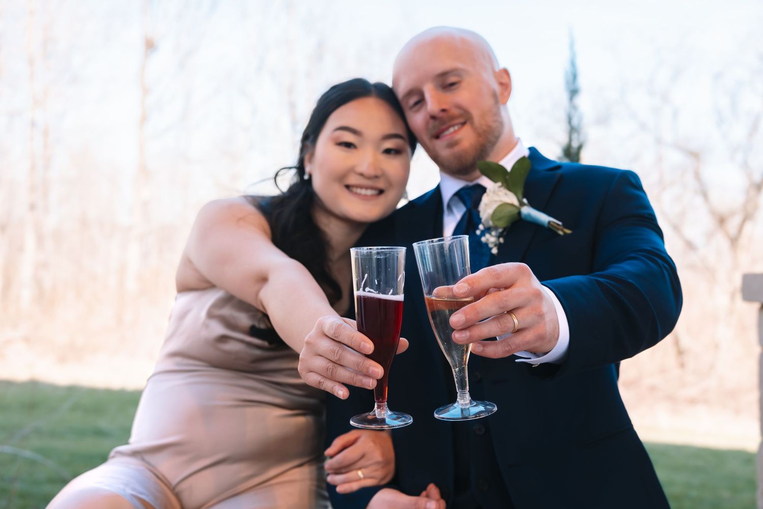 Smiling bride and groom in navy suit toasting with red champagne glasses at their wedding celebration.