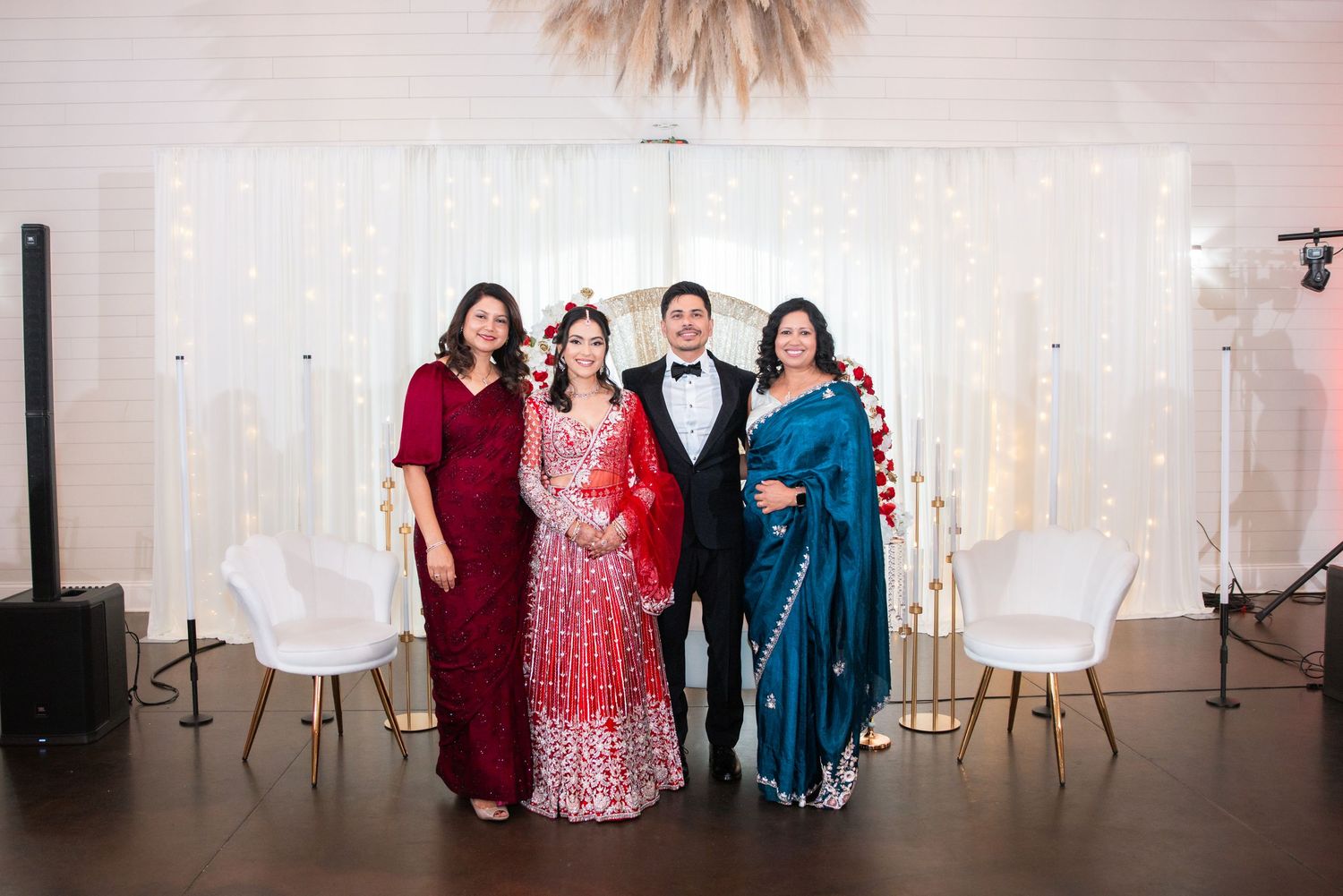 Indian wedding portrait of bride in red lehenga, groom in tuxedo, flanked by two women in elegant ethnic attire.