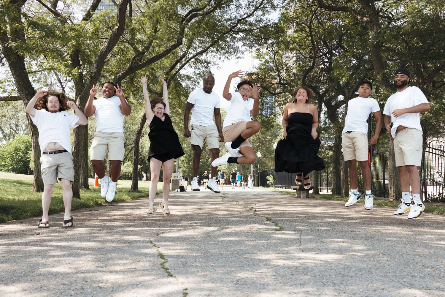 Joyful group of nine people jumping in the air together on a tree-lined park pathway.
