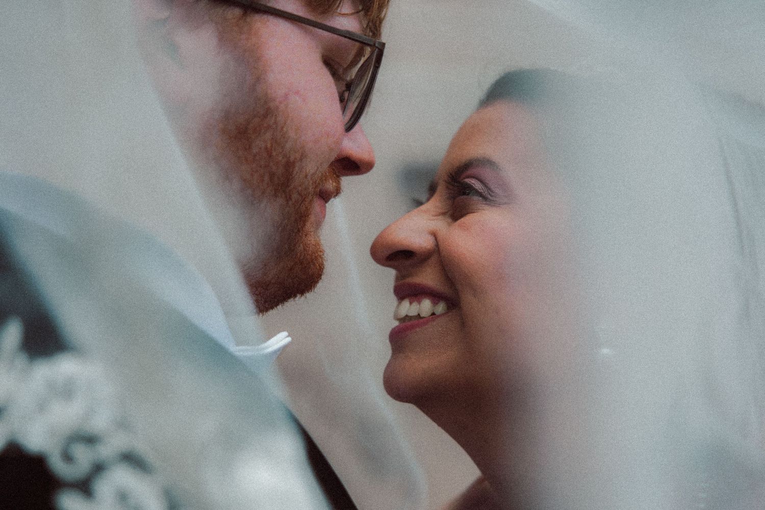 Bride smiling radiantly at groom under a lace-trimmed veil during wedding portraits.