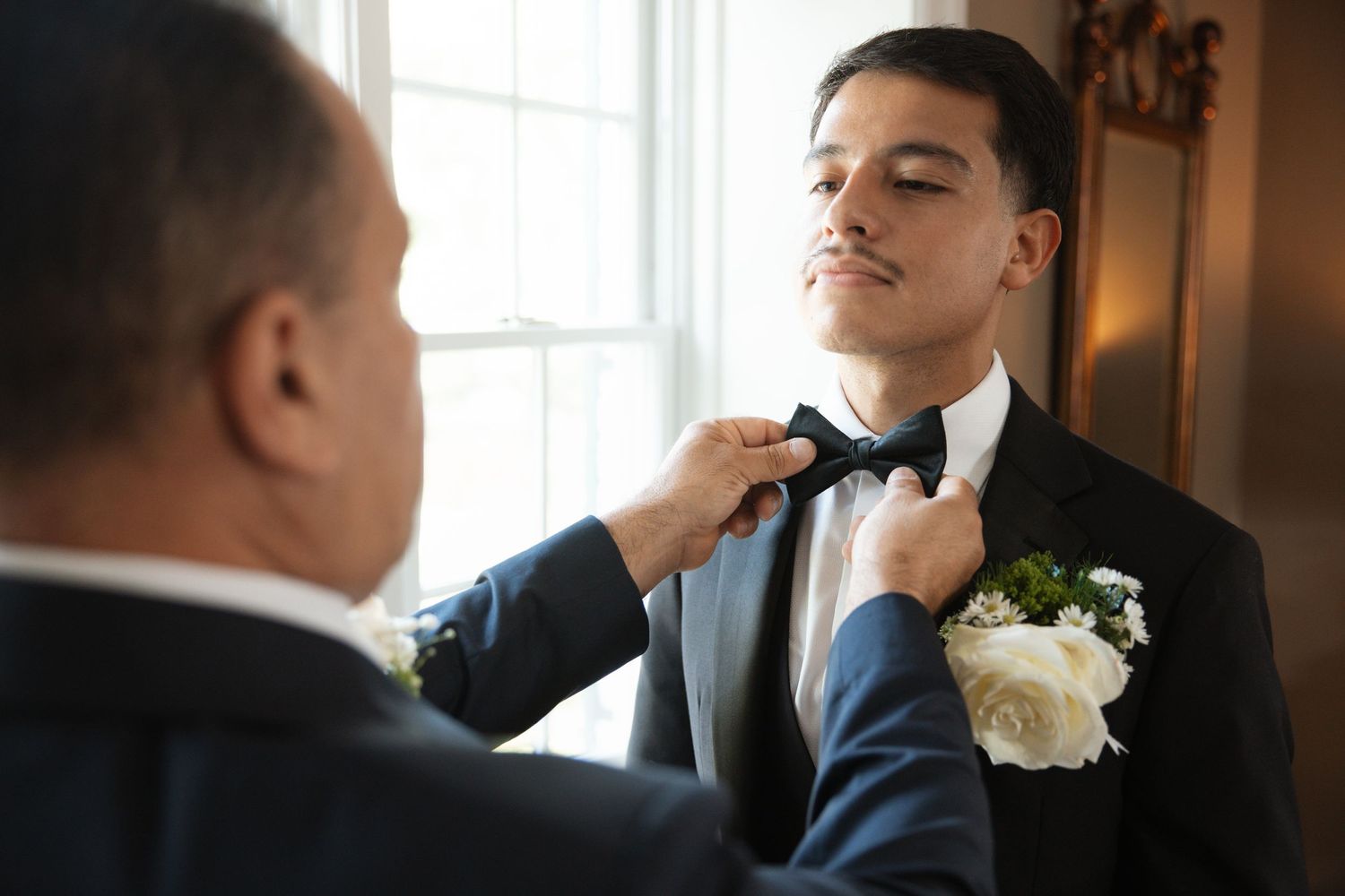 Man adjusting groom's black bow tie before wedding ceremony.