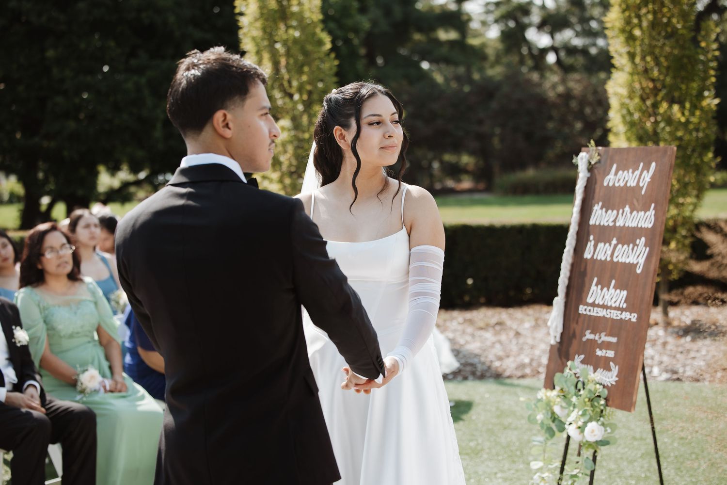 A bride in a white gown holds hands with the groom during an outdoor wedding ceremony.