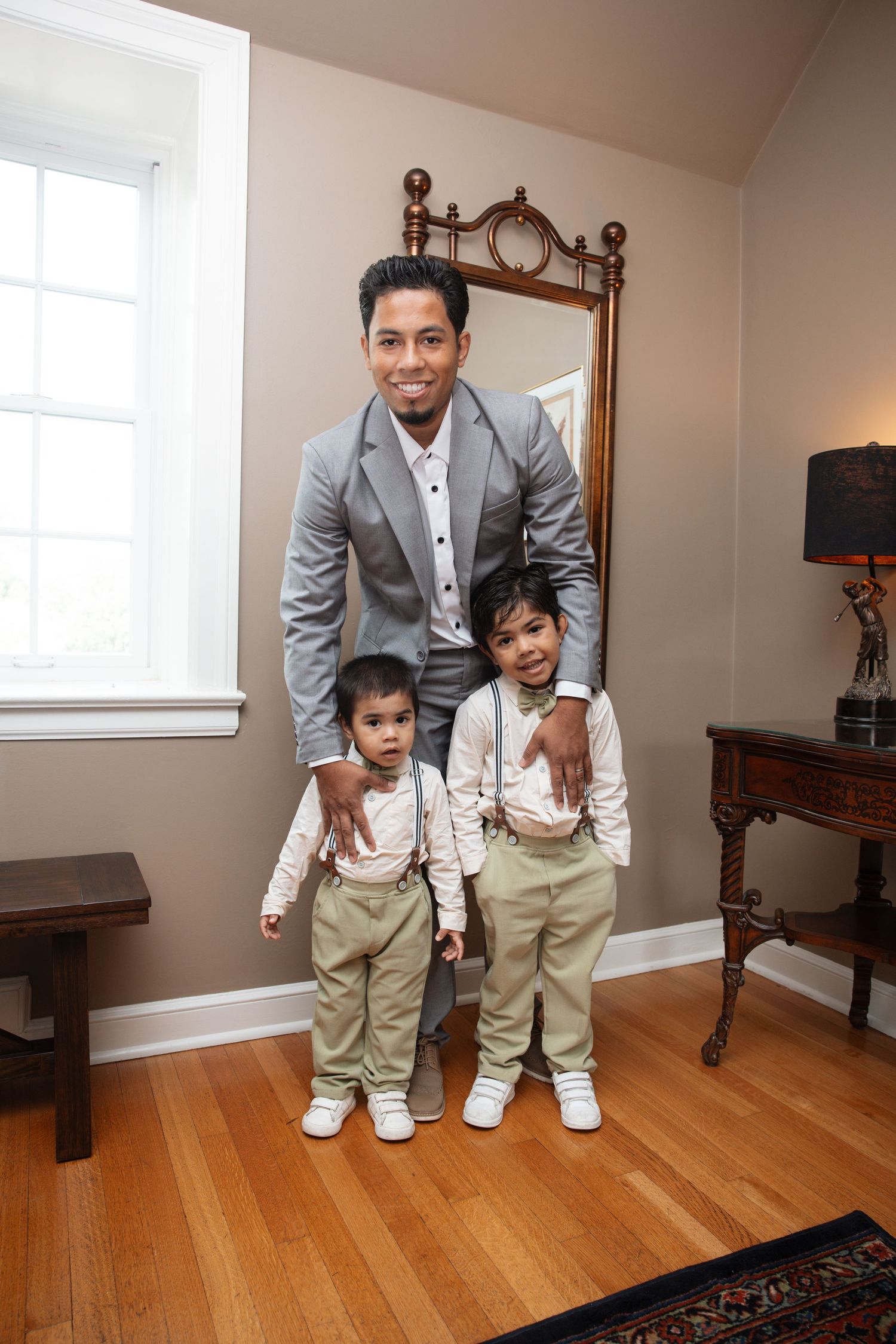 Man in gray suit posing with two young boys in matching khaki suspender outfits in a warmly decorated room.