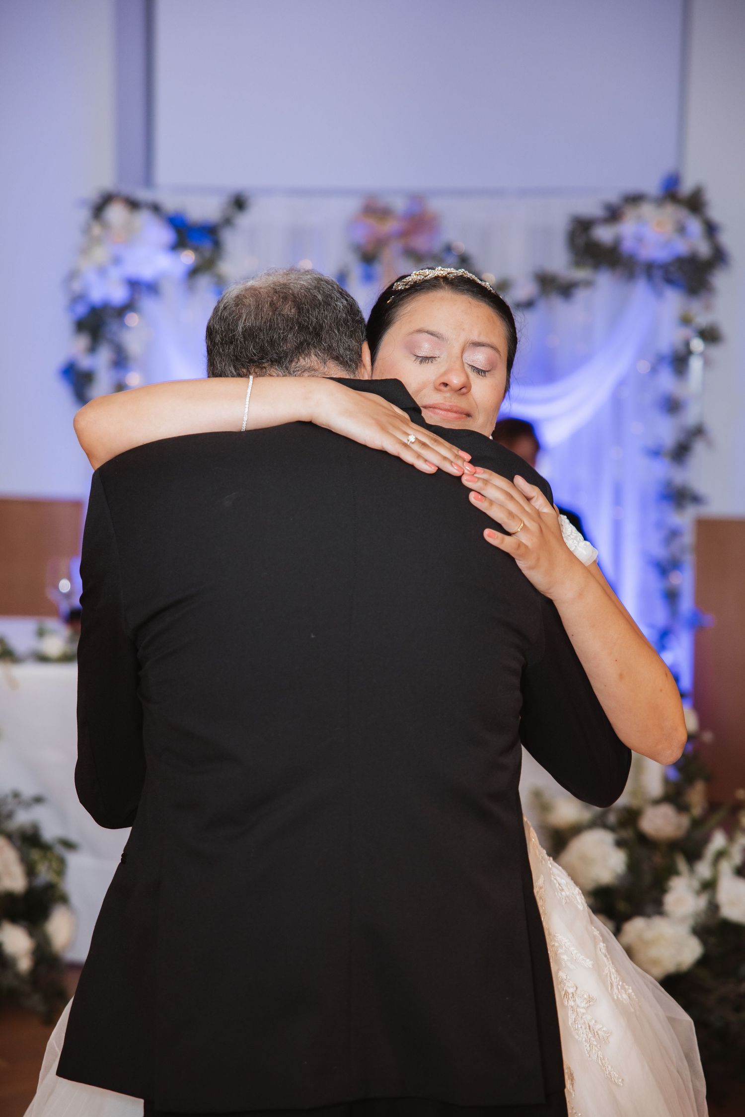 Emotional bride hugging partner during wedding dance with floral arch and blue decorations in background.