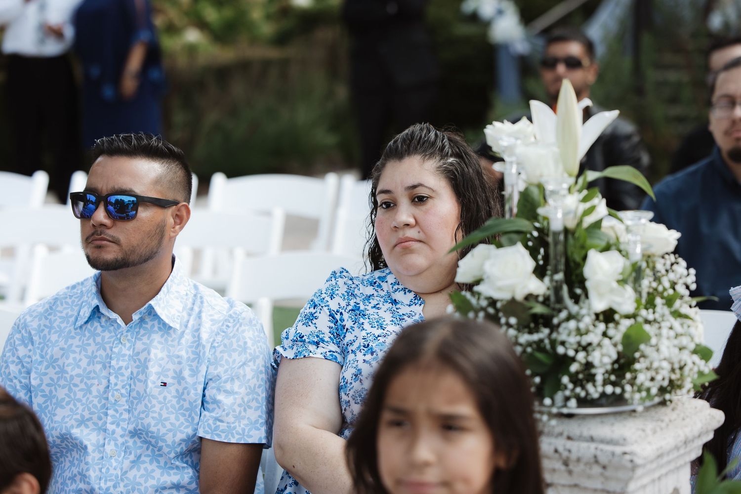 Couple in casual attire seated beside white floral arrangement at an outdoor wedding ceremony.