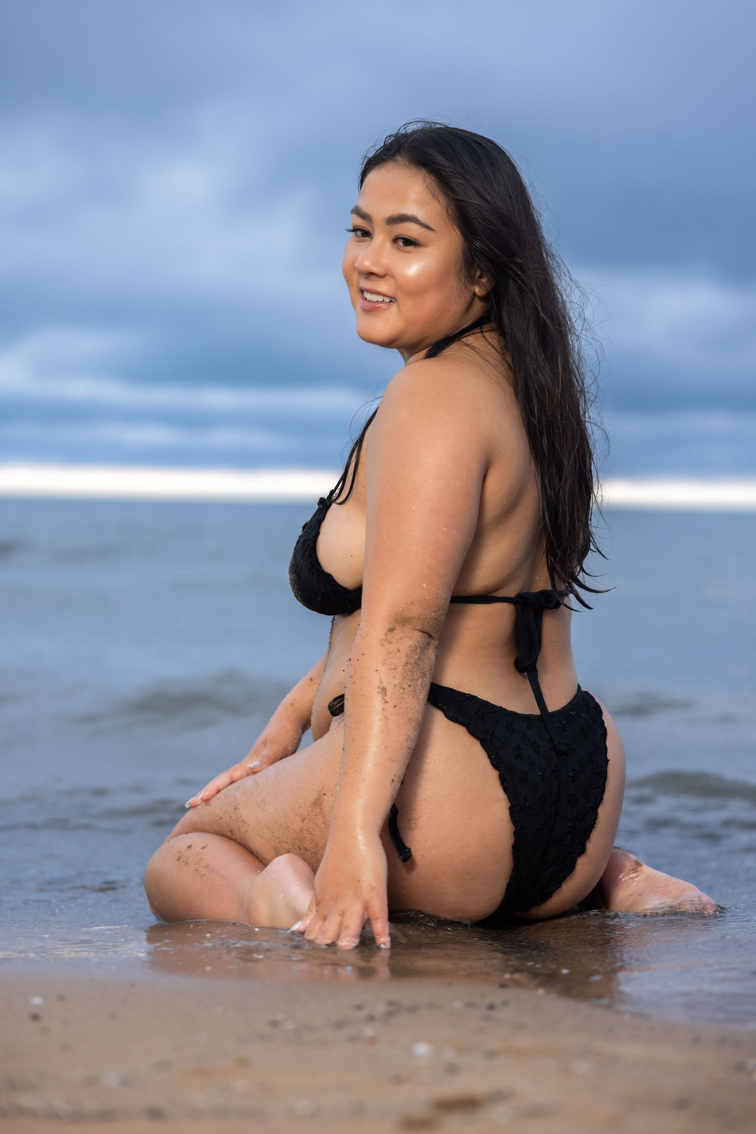 Smiling woman in black bikini sitting on wet sand at beach with ocean waves in background.