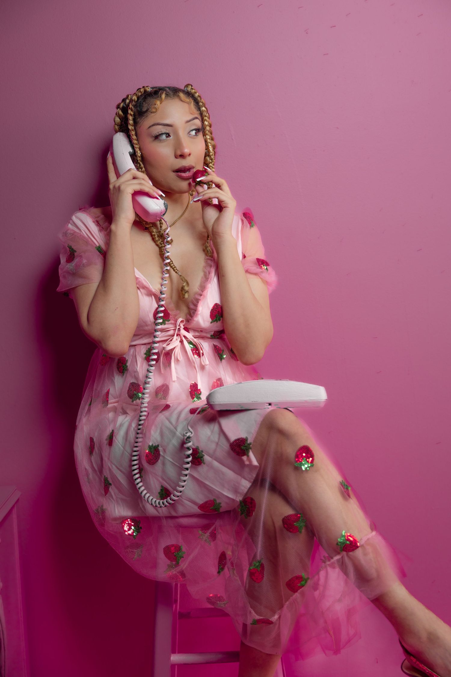 Woman in pink strawberry dress talks on vintage corded phone against pink wall.