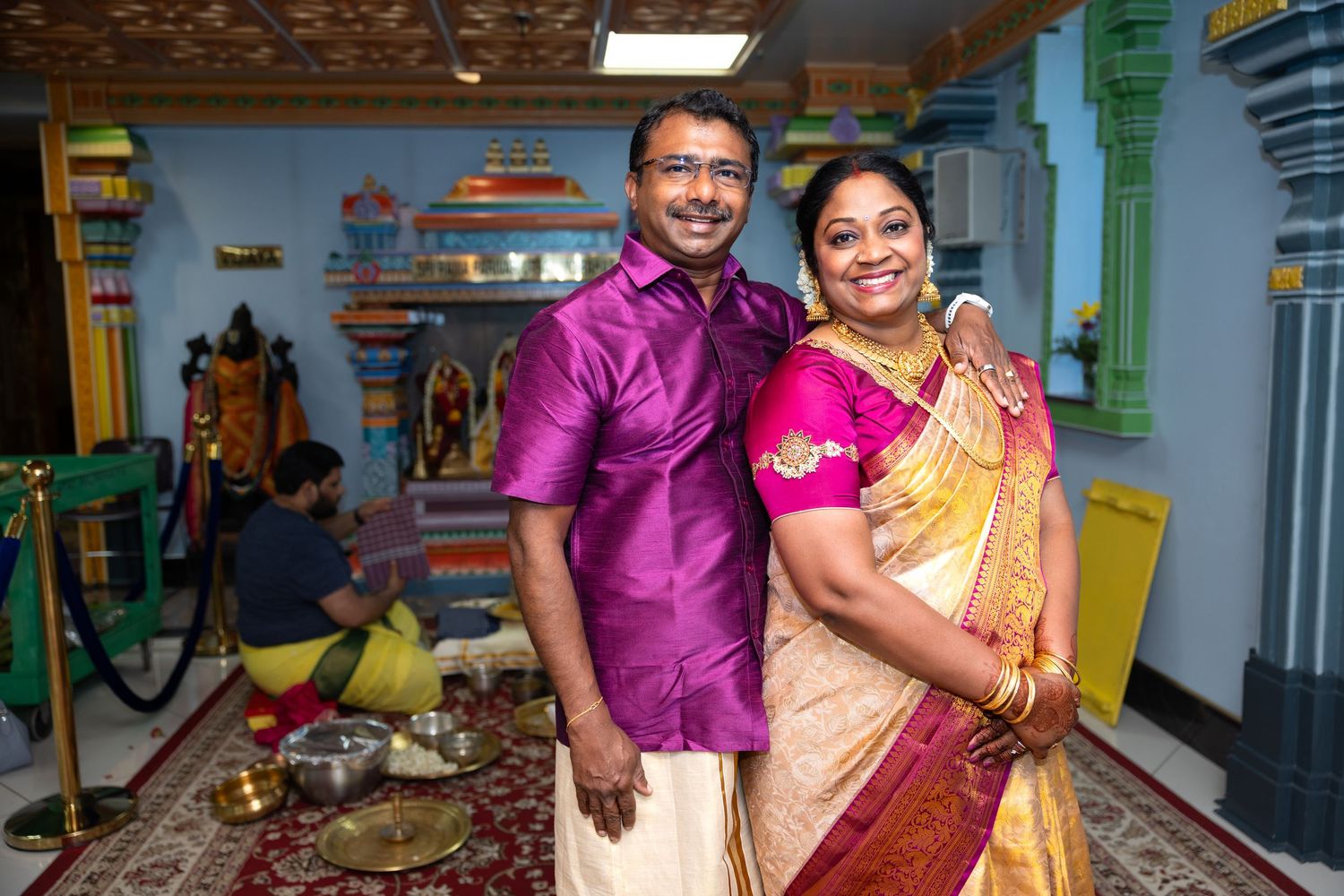 Smiling Indian couple in traditional purple and gold festive attire pose inside ornate temple.