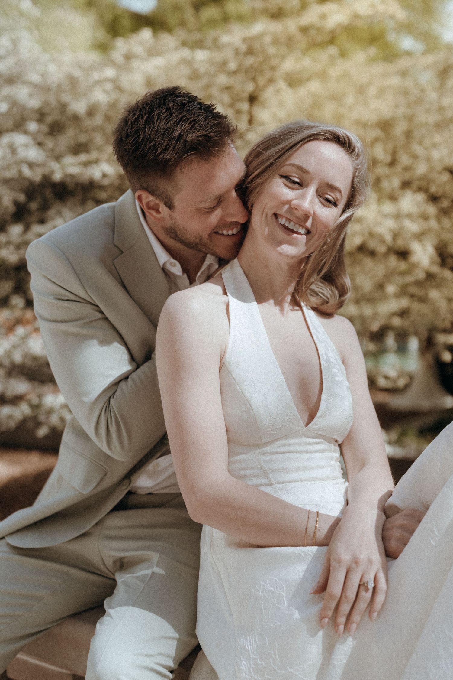 Laughing bride in white halter gown embraced from behind by smiling groom in a sage suit outdoors.