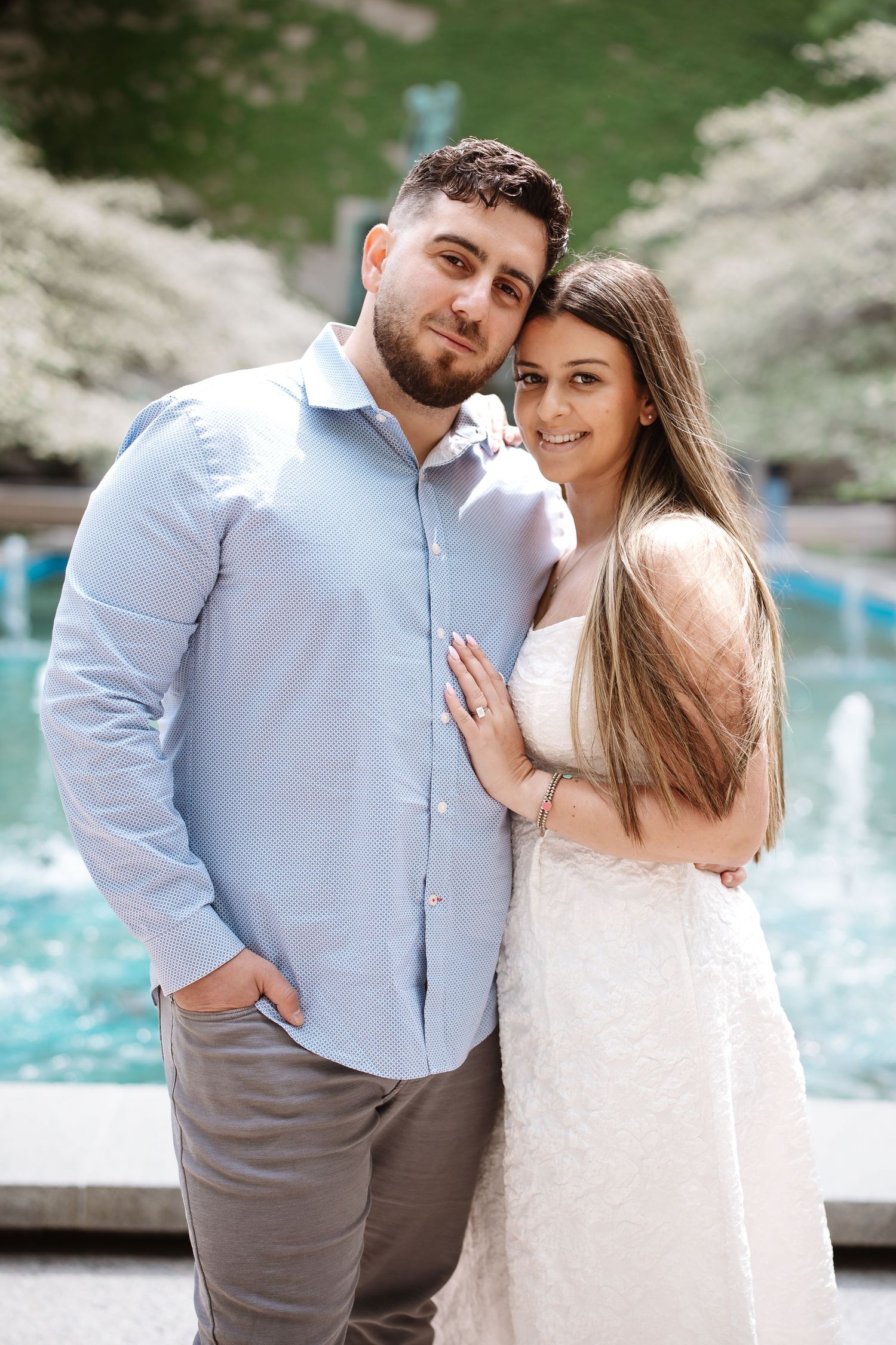 Couple posing by a fountain outdoors, woman in white dress leaning on man in blue shirt, smiling at camera.