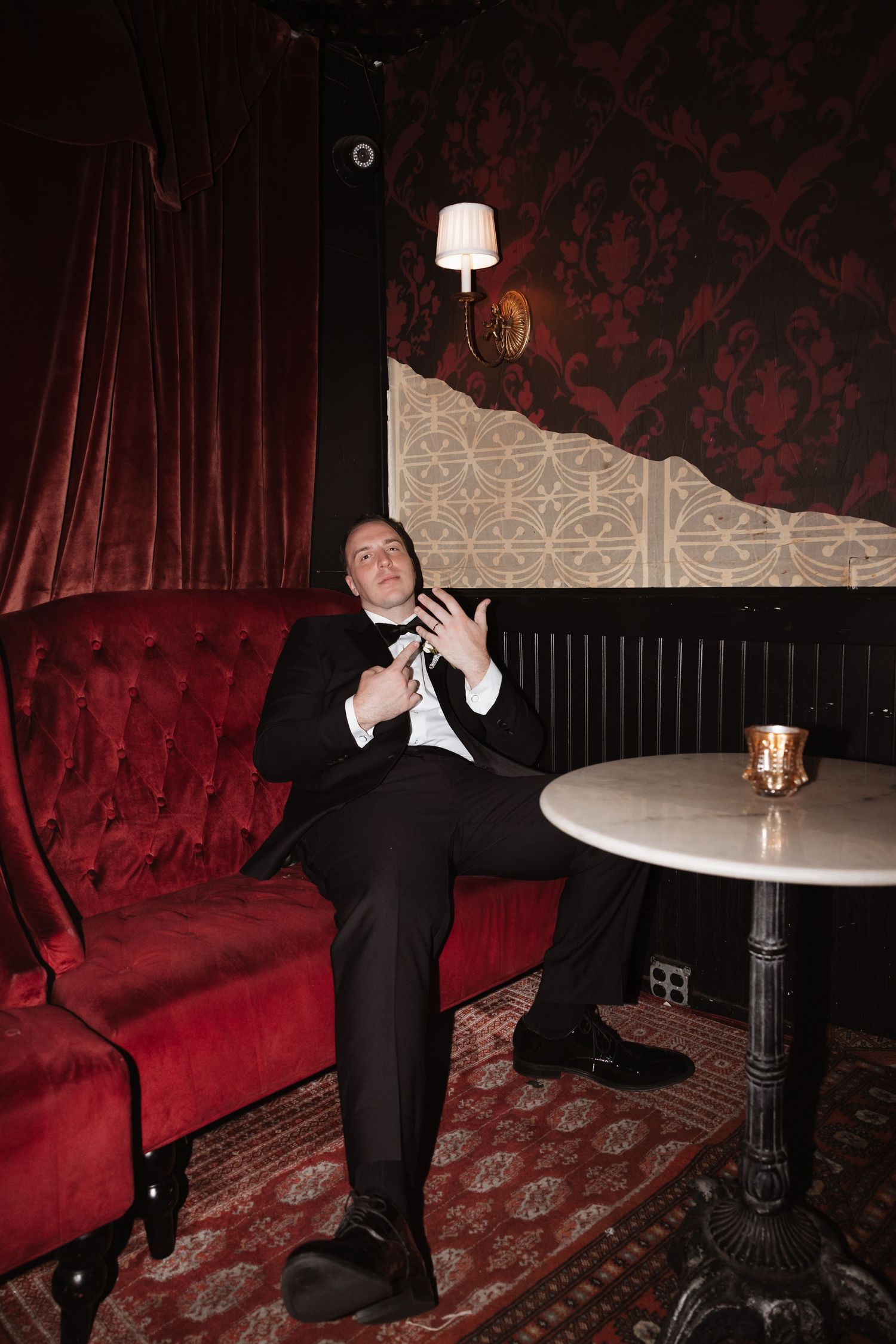 A man in a tuxedo sits on a red velvet sofa in an elegant lounge with damask walls and a marble side table.