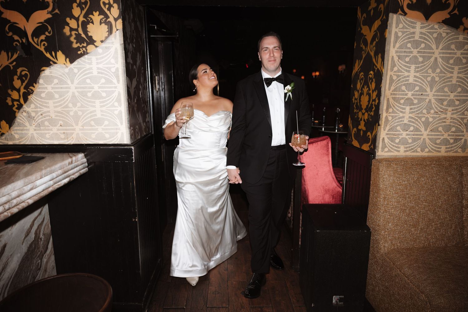 Bride and groom enter their wedding reception holding champagne glasses in a dark elegant venue.