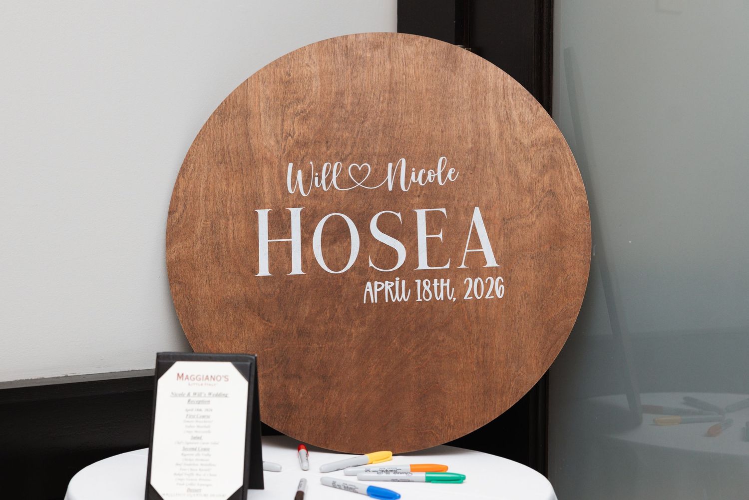 Round wooden wedding sign for Will and Nicole Hosea dated April 13th, 2026 on a white table.