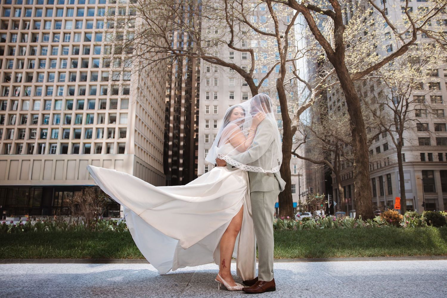 Bride and groom embrace outdoors with veil flowing in wind, city buildings and spring trees behind.