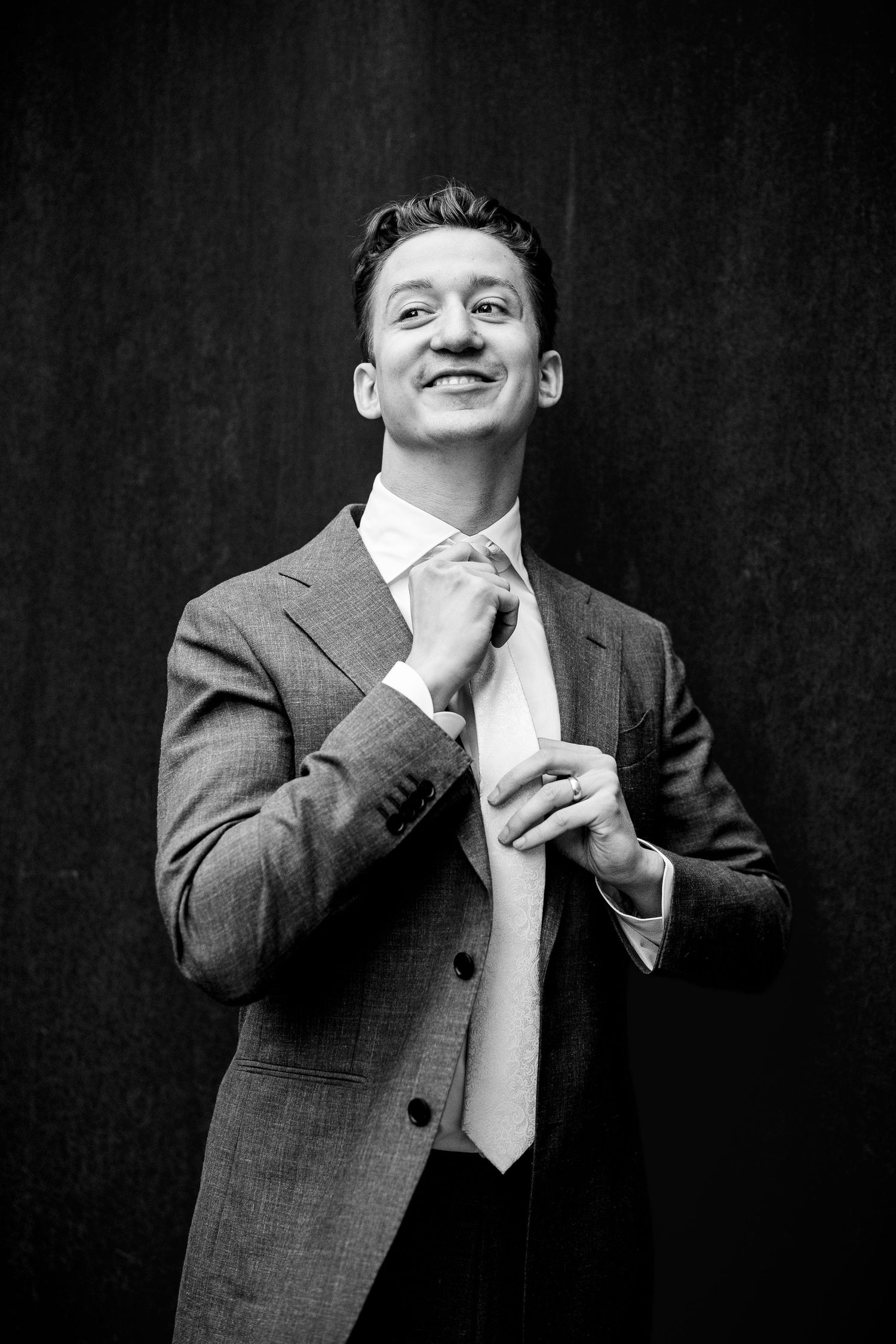 Smiling groom adjusts tie in black and white portrait photo while wearing a classic gray wedding suit.