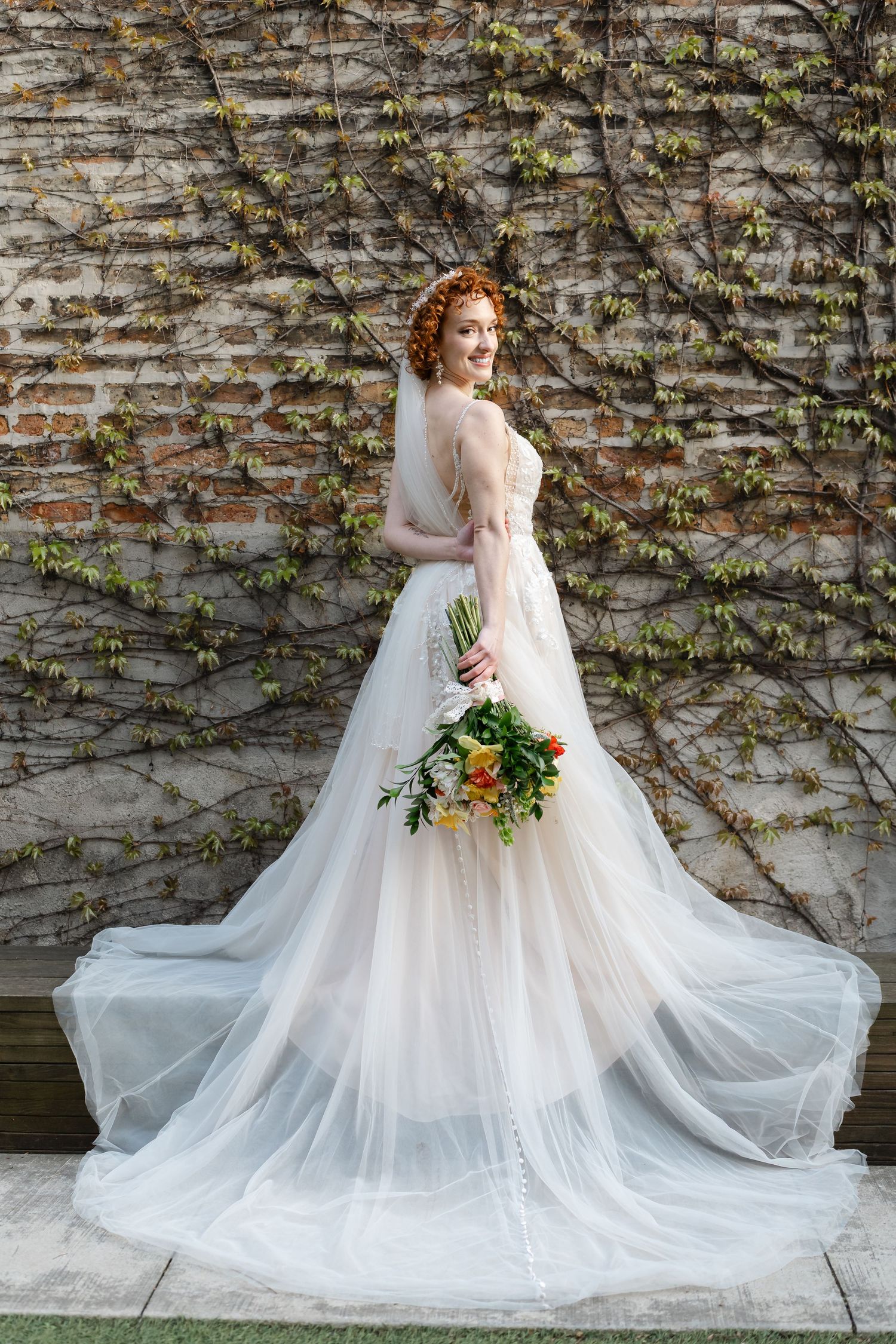Elegant bride in a flowing white tulle gown holding a colorful bouquet, posing before an ivy-covered brick wall.
