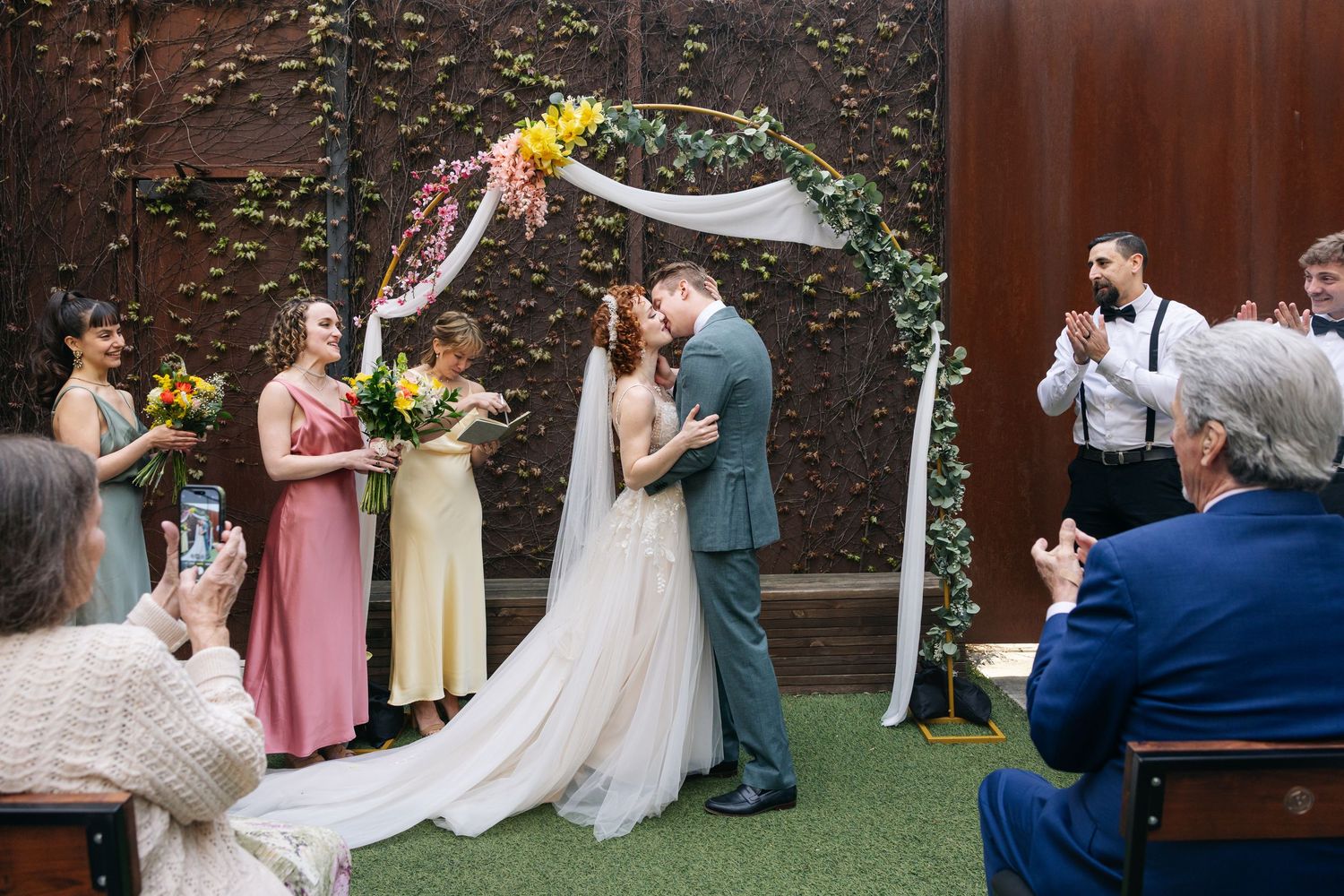 Bride and groom share first kiss under floral arch while wedding party and guests applaud at outdoor ceremony.