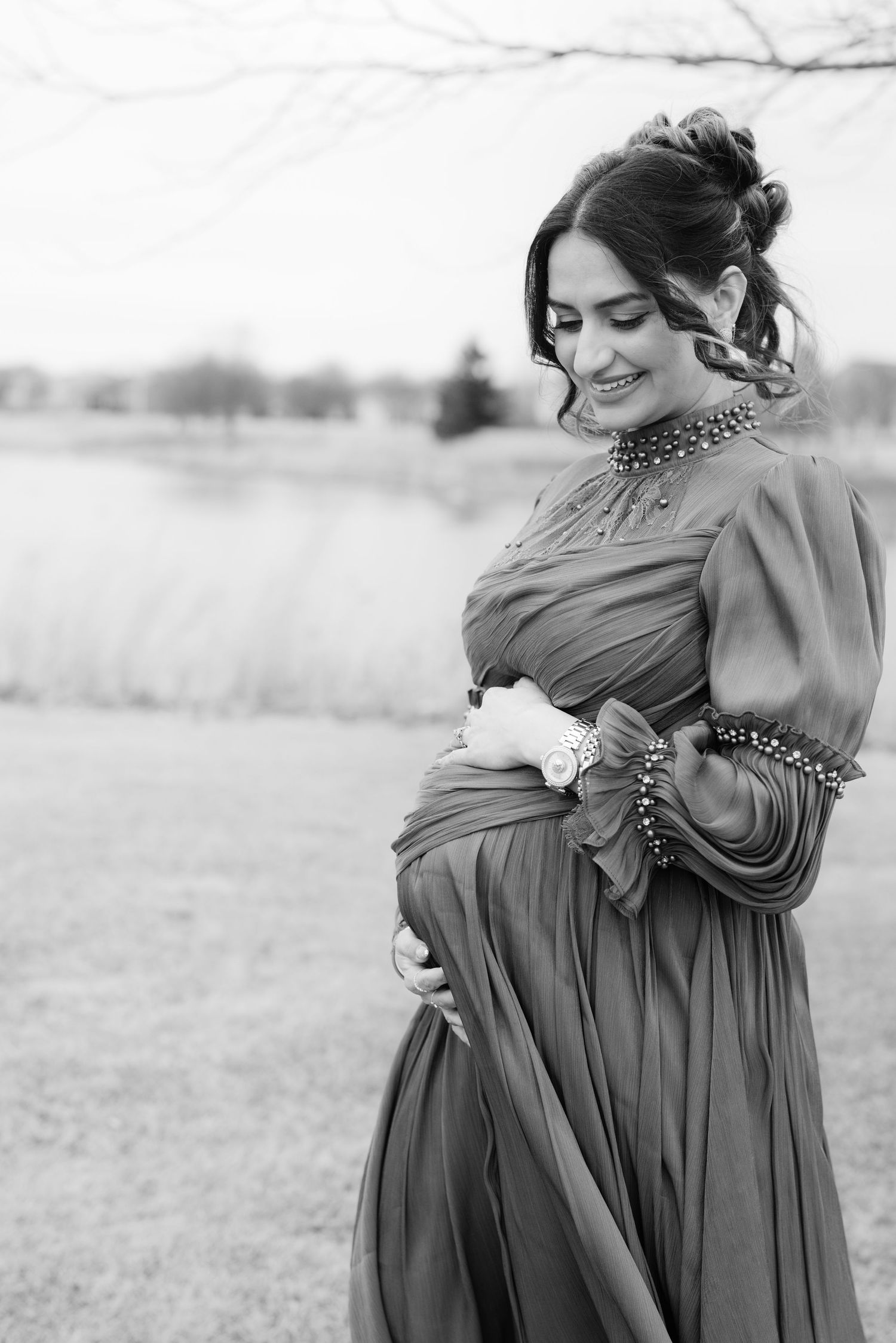 Black and white portrait of pregnant woman in elegant gown cradling baby bump near a lake.