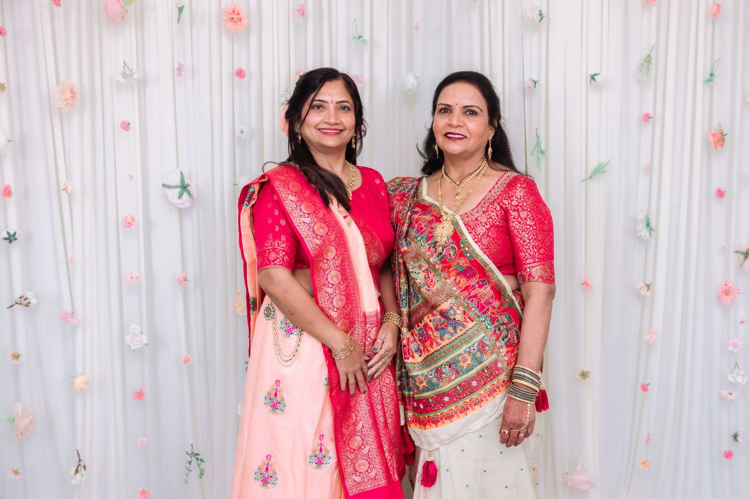 Two Indian women in vibrant pink traditional sarees and salwar suits smiling together at a festive celebration.