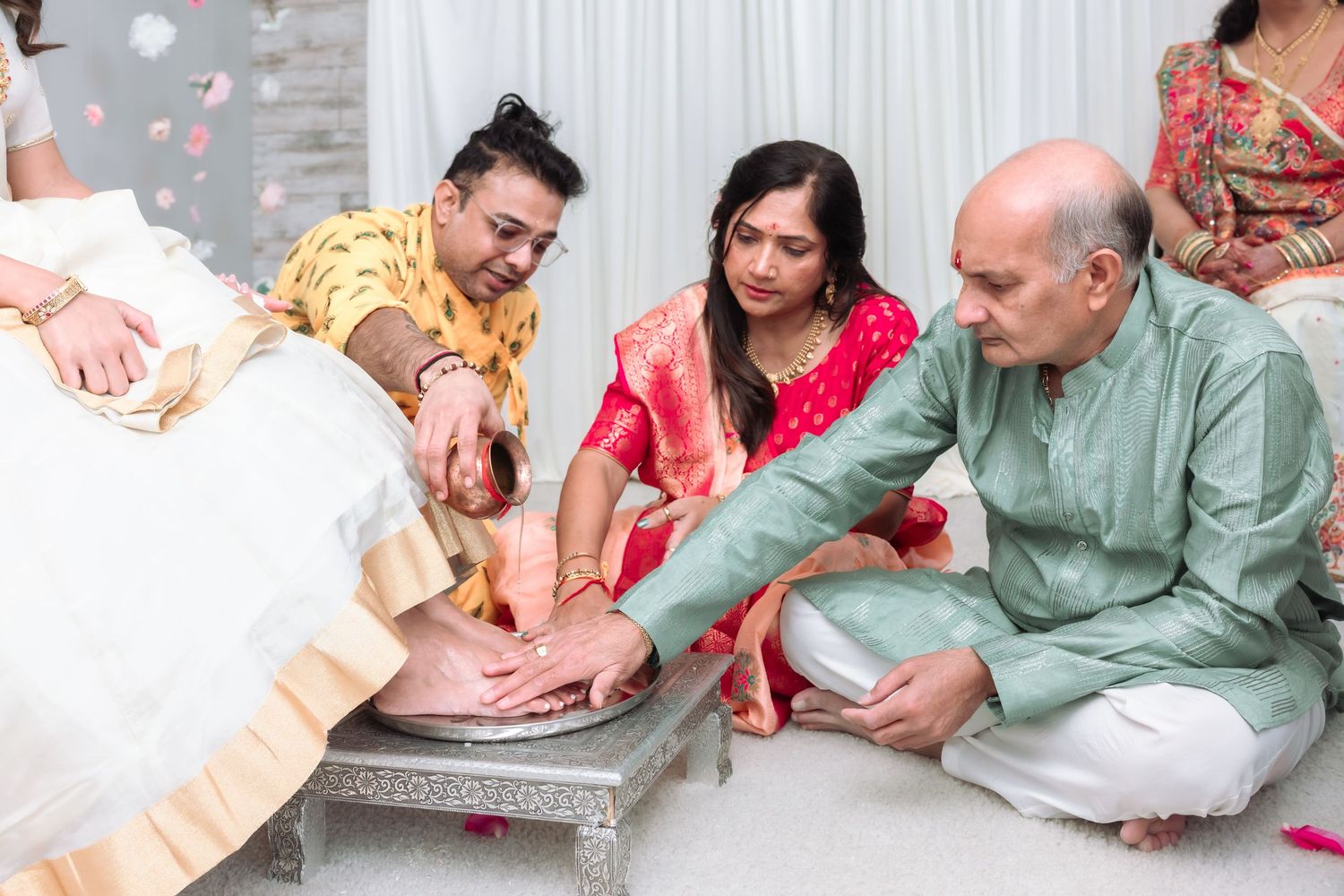 Family members in traditional Indian attire perform a ceremonial foot washing ritual during a wedding.