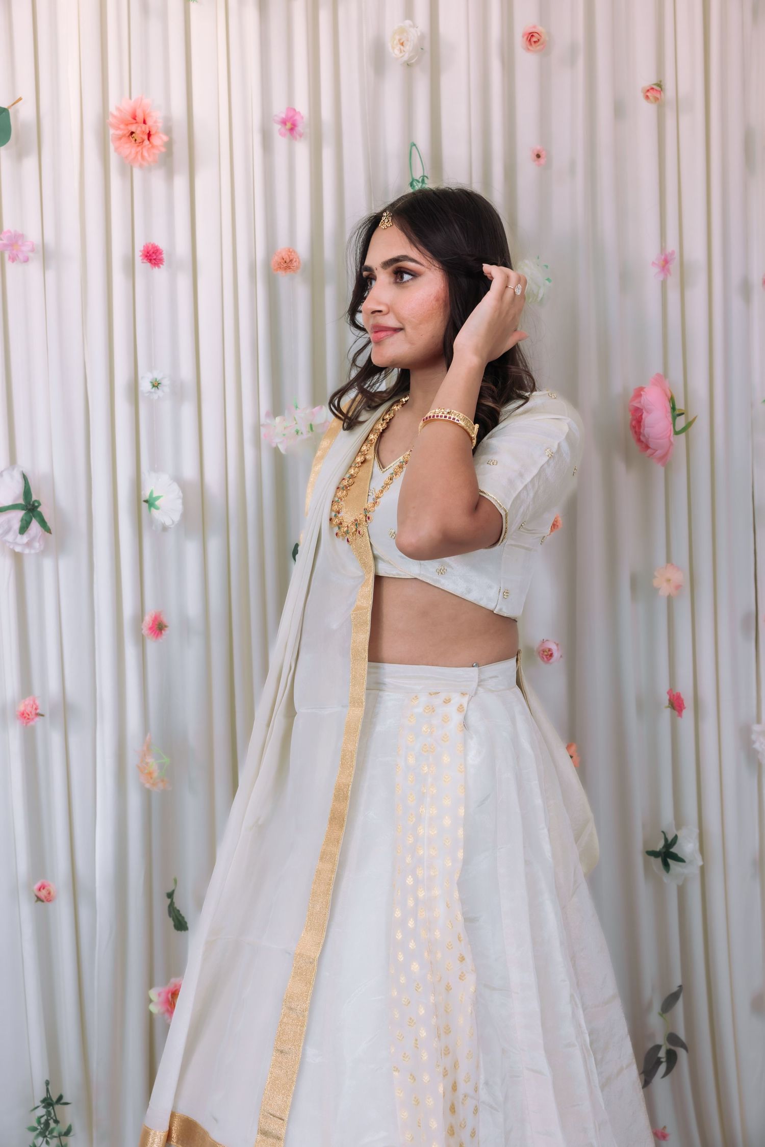 Woman in white and gold lehenga with dupatta posing before floral curtain backdrop.