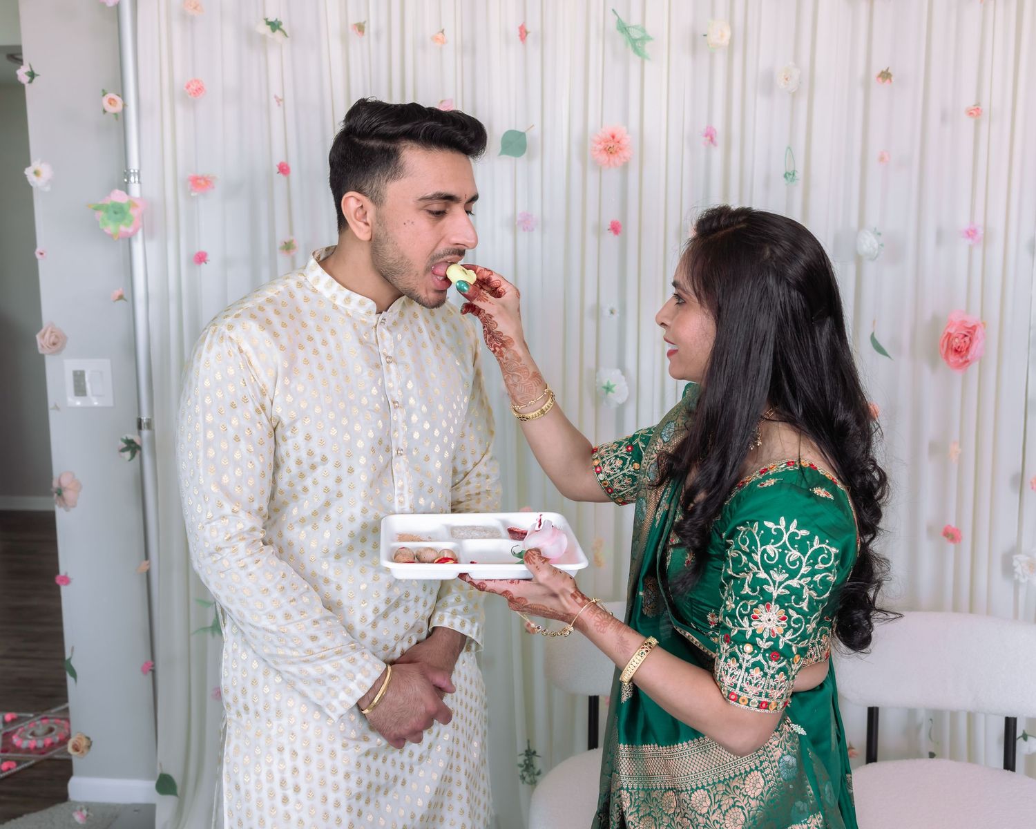 A woman in green lehenga feeds sweets to a man in white sherwani at an Indian engagement ceremony.