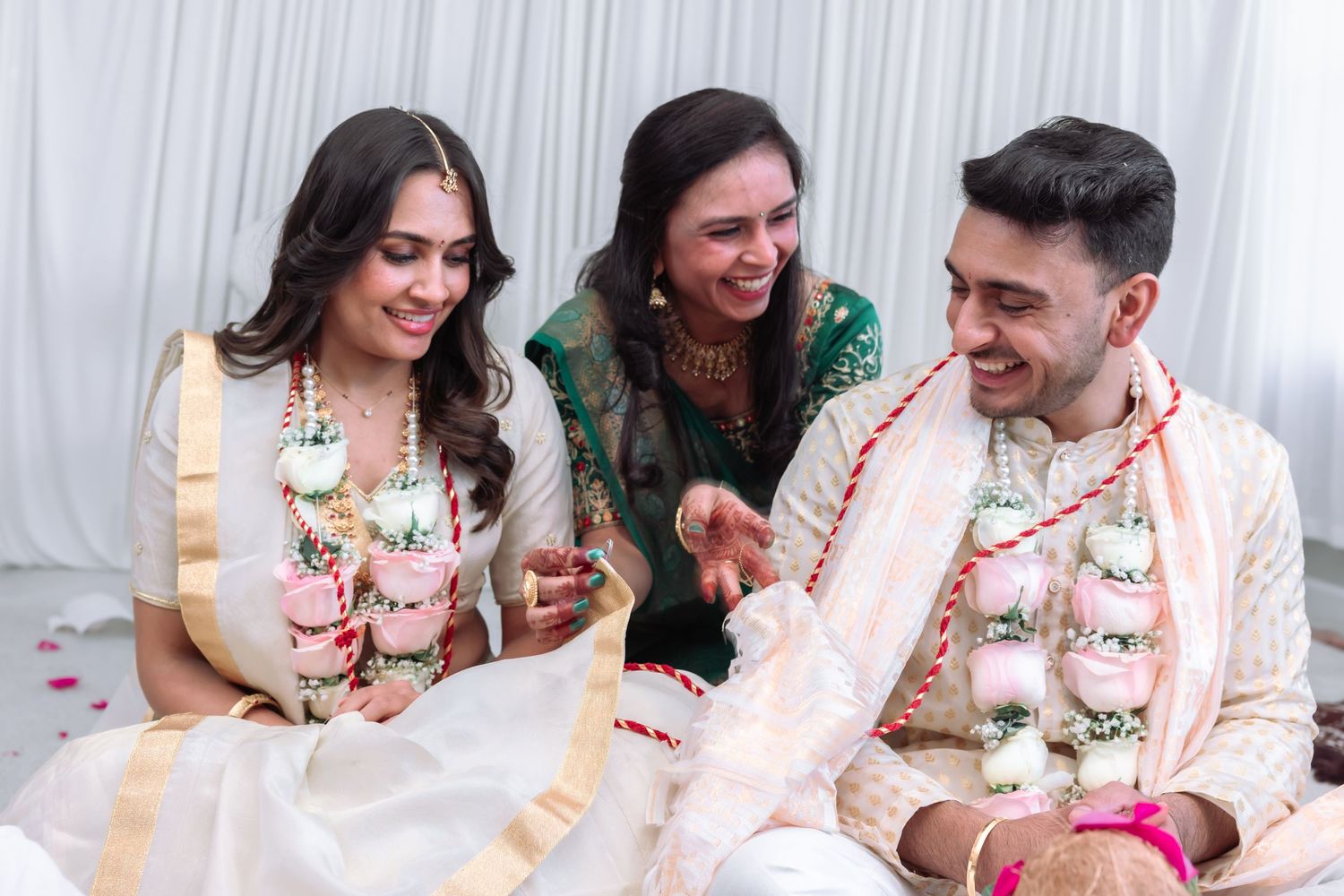 Bride and groom in traditional Indian wedding garlands share a joyful moment with a female guest.