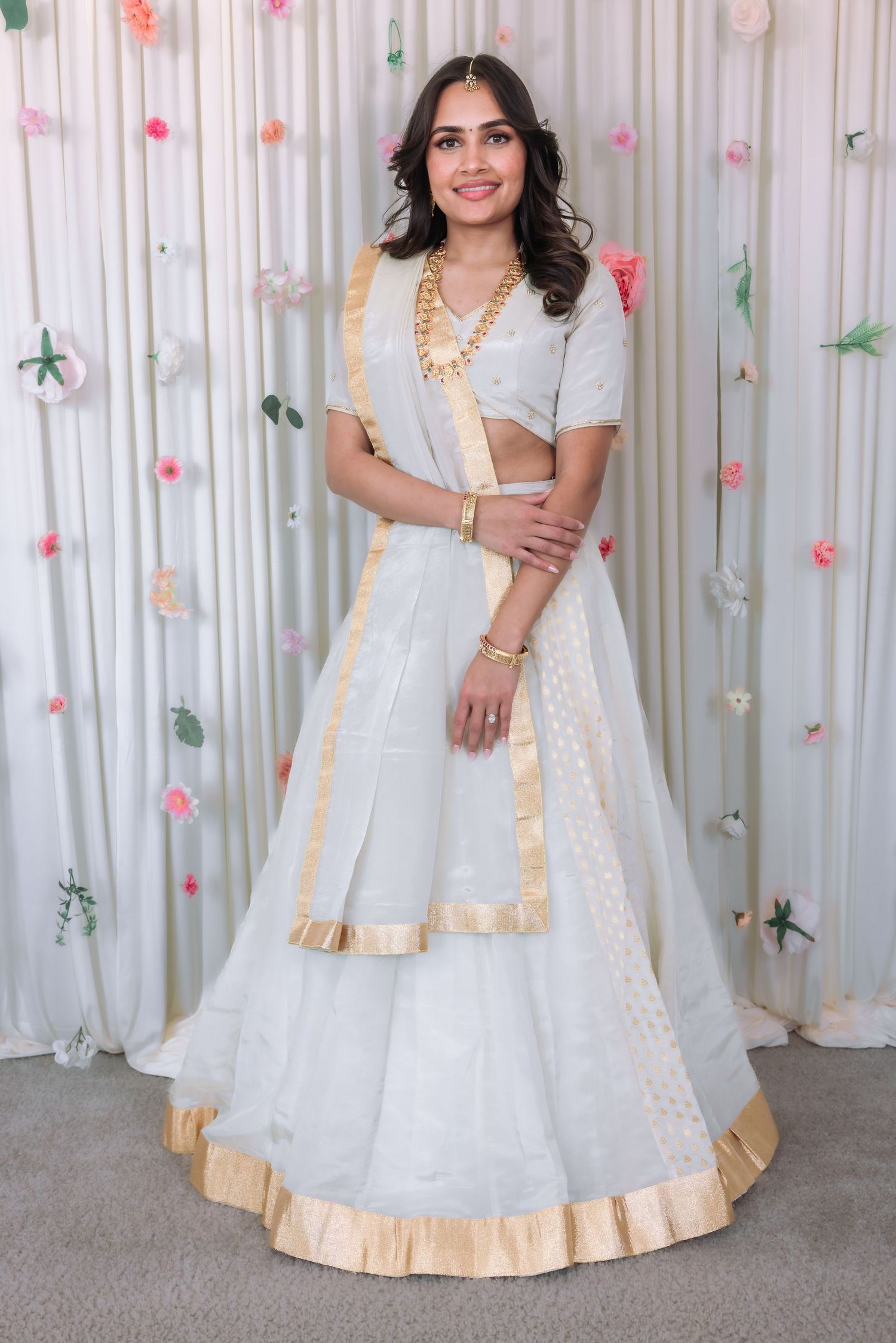 Woman wearing white lehenga with gold trim and maang tikka smiling before a floral backdrop.