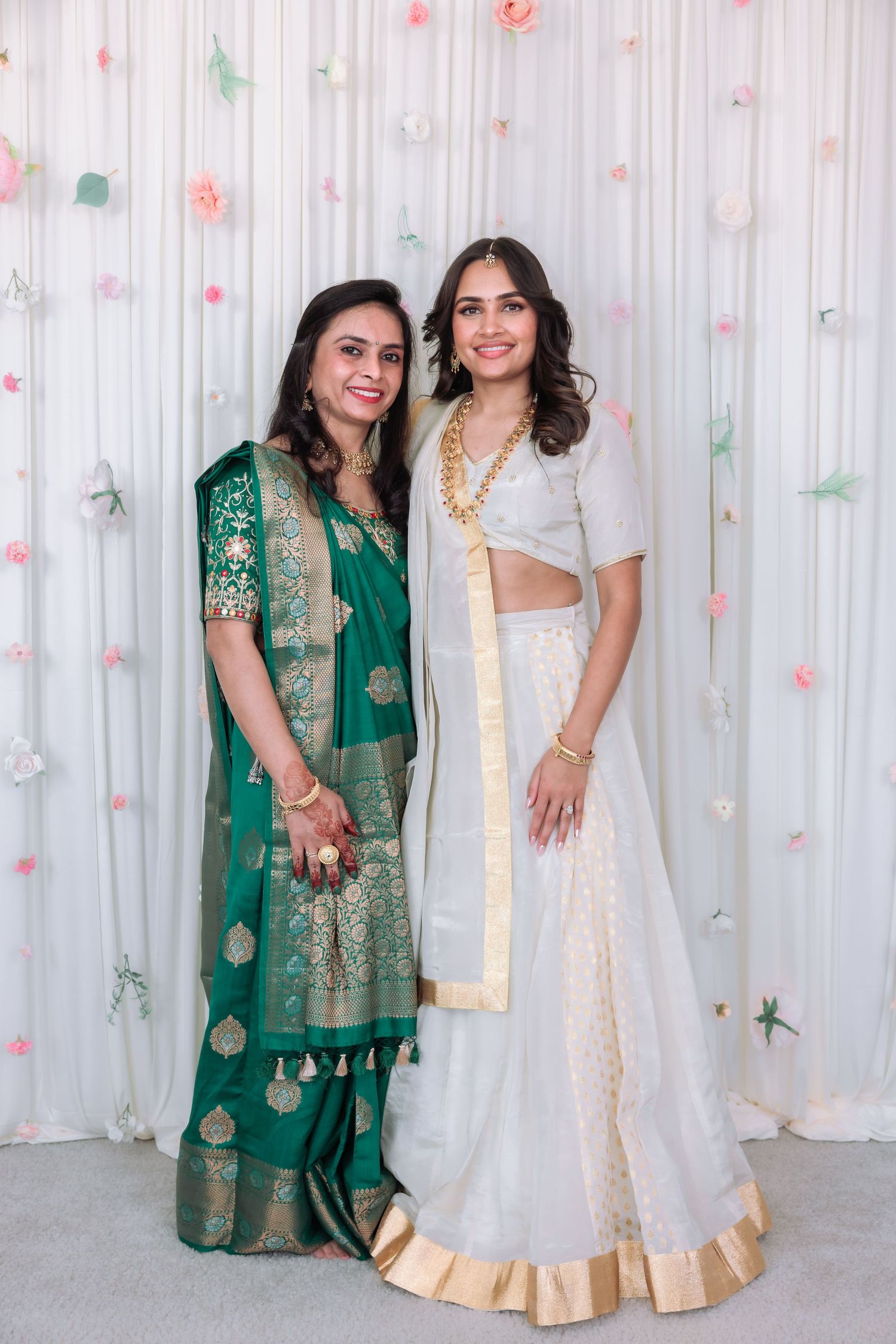 Indian bride in white lehenga poses with older woman in green saree before floral wedding backdrop.