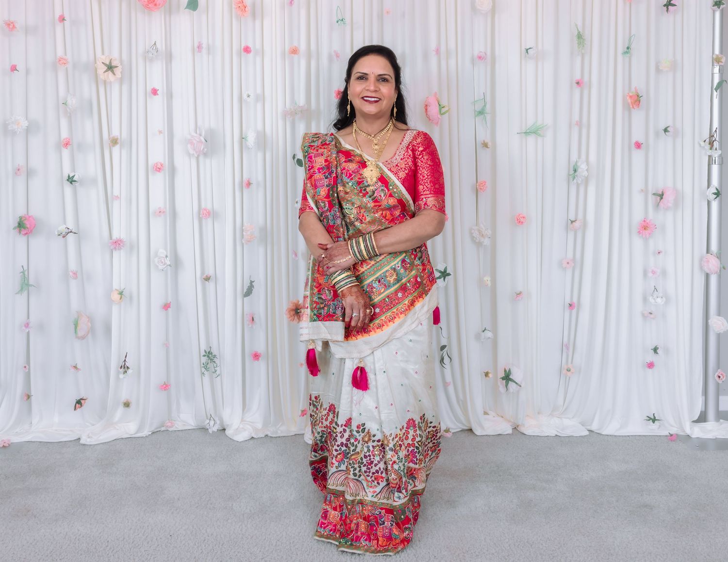 Indian woman in elegant white and red embroidered lehenga with gold jewelry poses at floral decorated wedding backdrop.