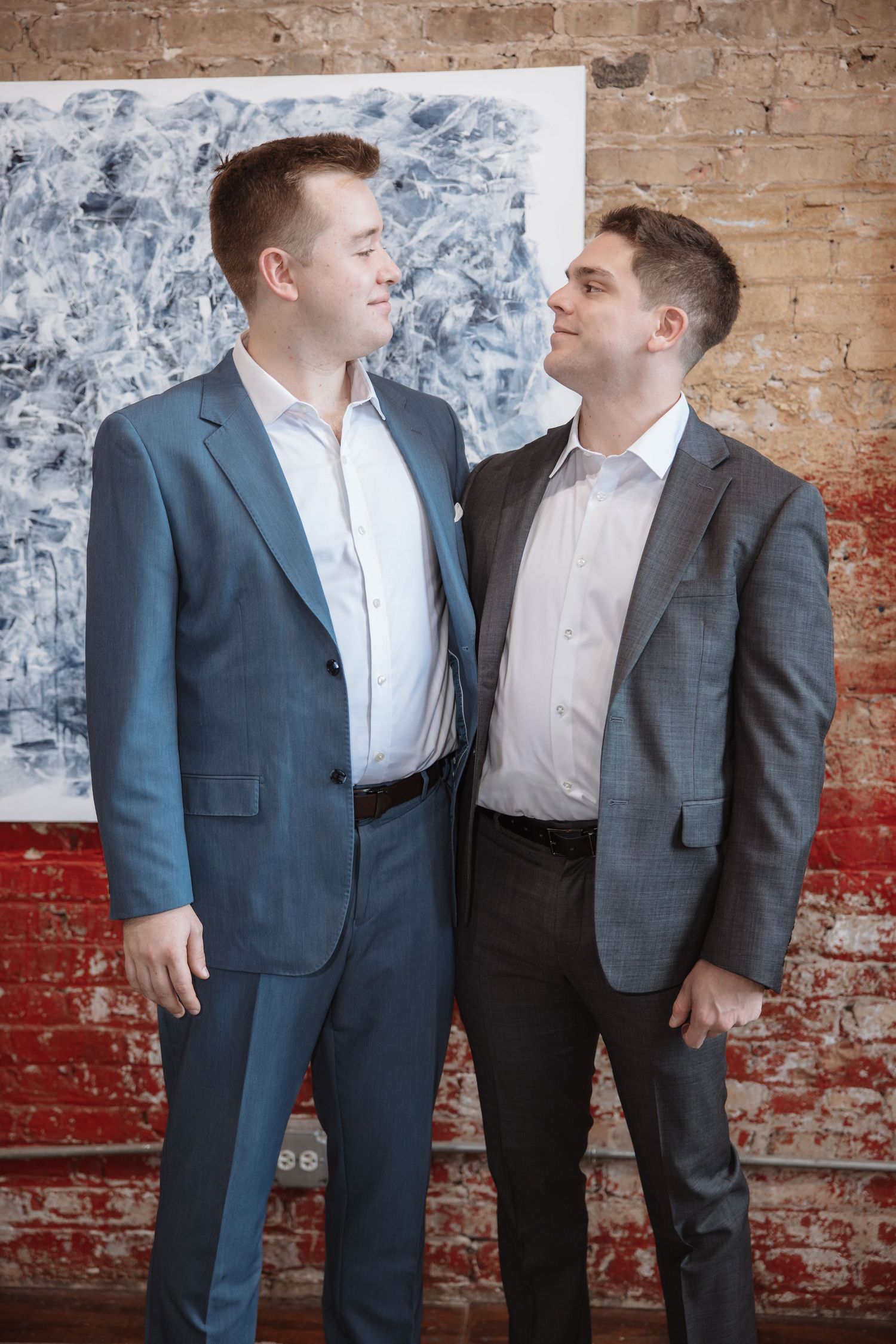 Two men in suits smile at each other in front of abstract art on a brick wall.