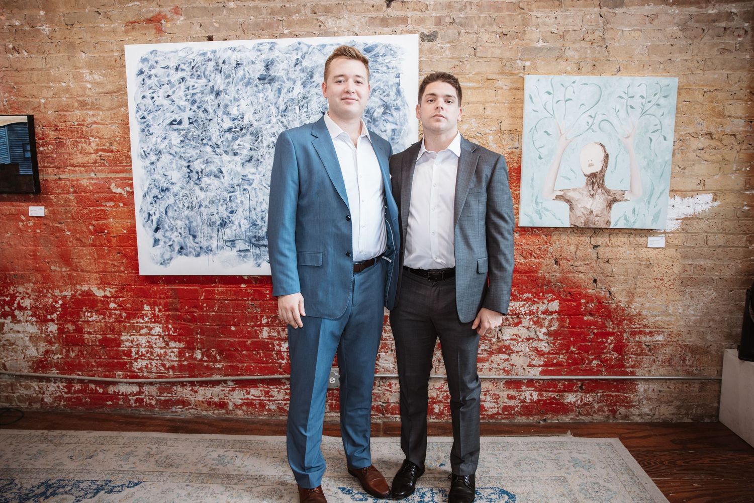 Two men in suits stand together in an art gallery with exposed brick walls and paintings.
