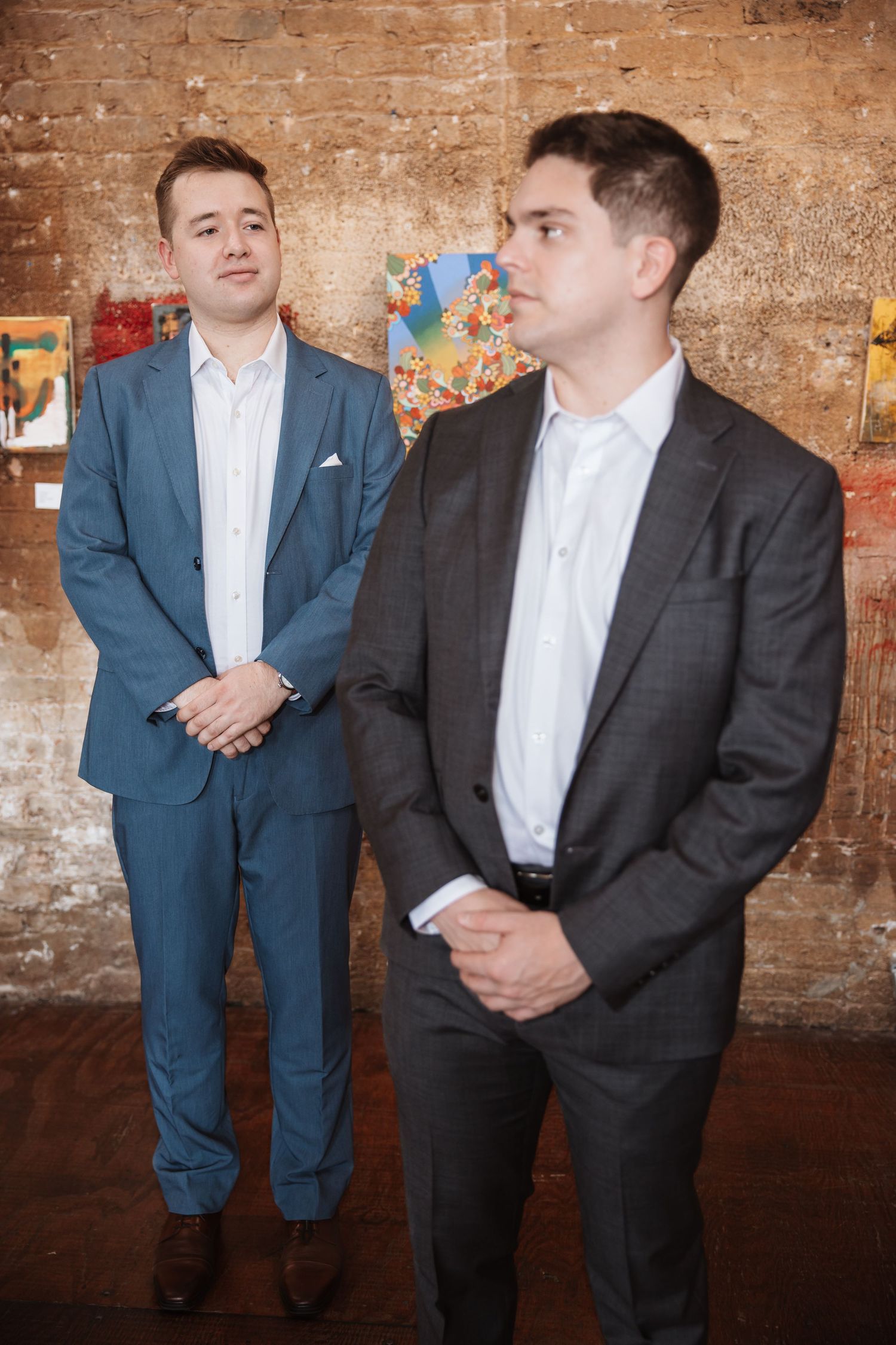 Two men in suits stand attentively at an art gallery event surrounded by colorful artwork.