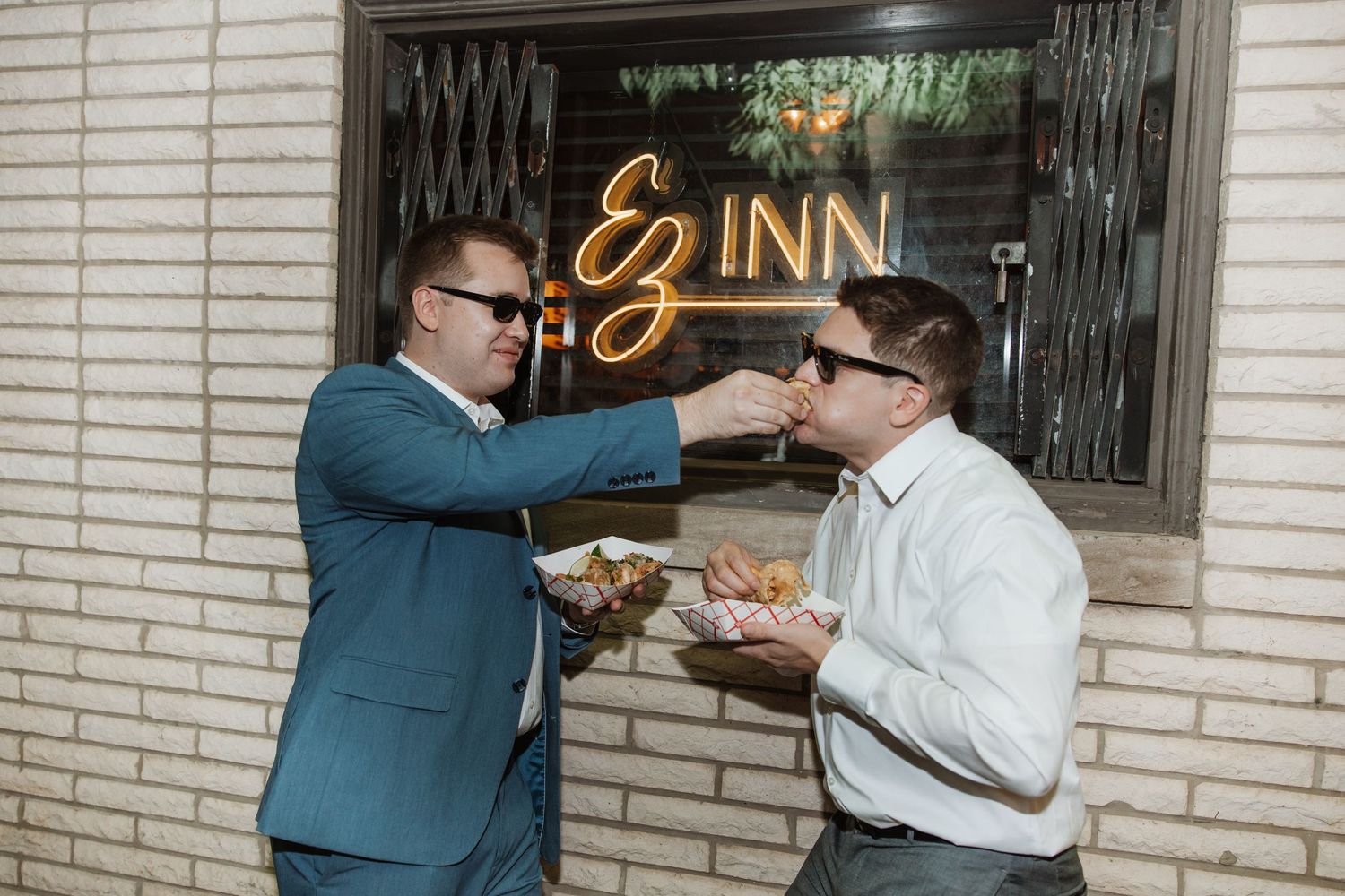 Two men in suits feeding each other food outside the EZ Inn neon sign entrance.