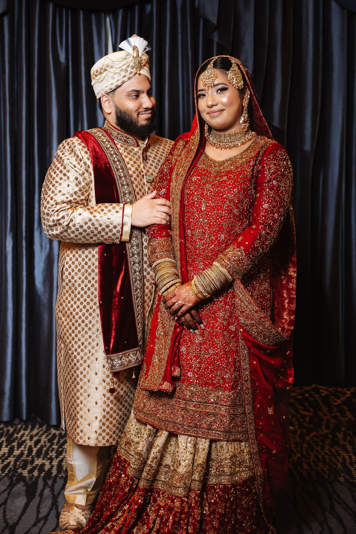 Pakistani bride in red embroidered bridal dress with groom in gold sherwani posing for wedding portrait.