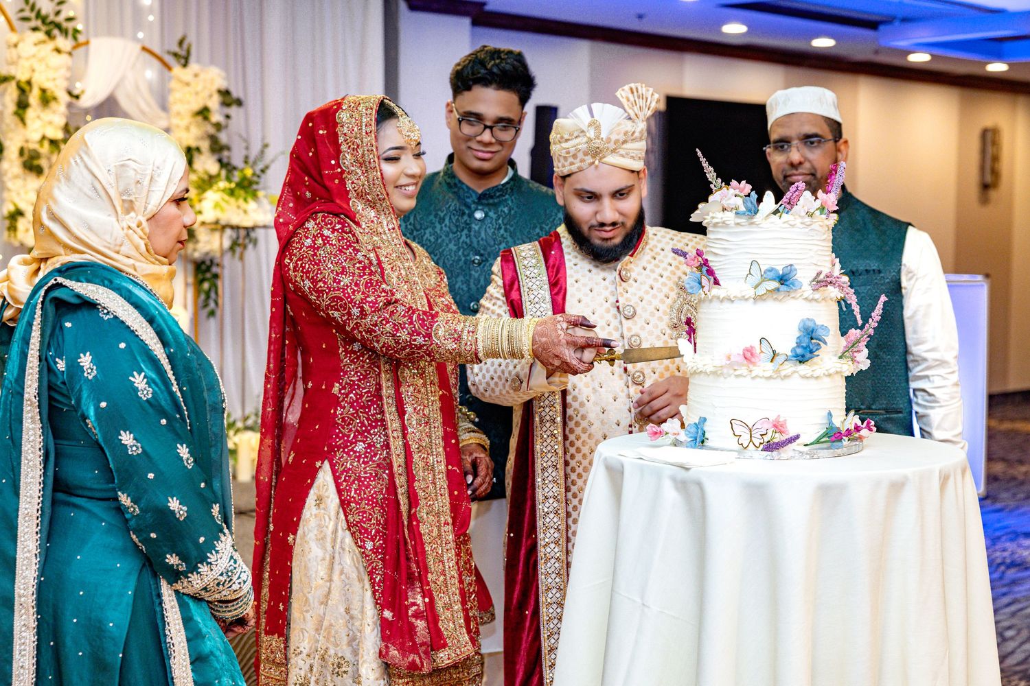Bride and groom in traditional South Asian attire cutting wedding cake with guests.