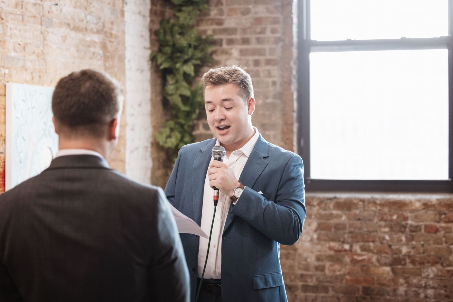 Young man in blue suit speaks into microphone at an elegant indoor event.