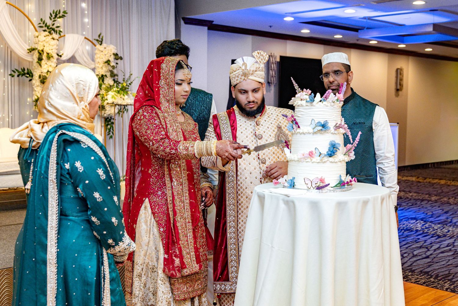 South Asian bride and groom cut butterfly-decorated wedding cake with family at reception.