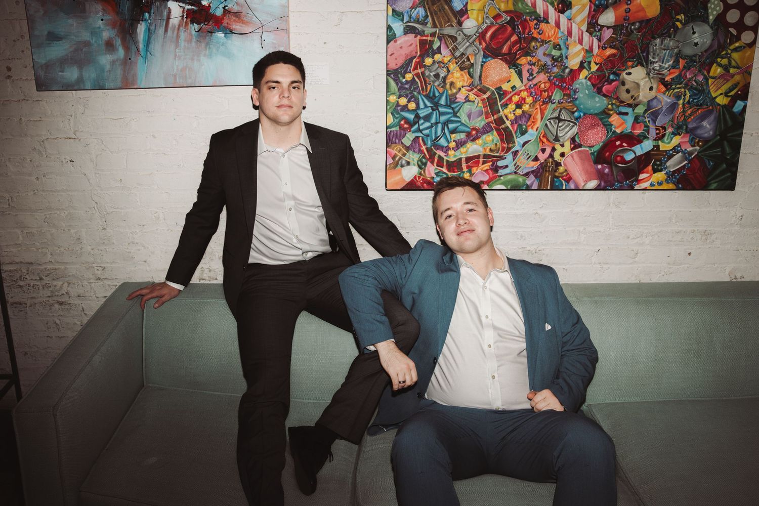 Two stylish men in formal suits sitting and leaning against a wall with vibrant abstract paintings.