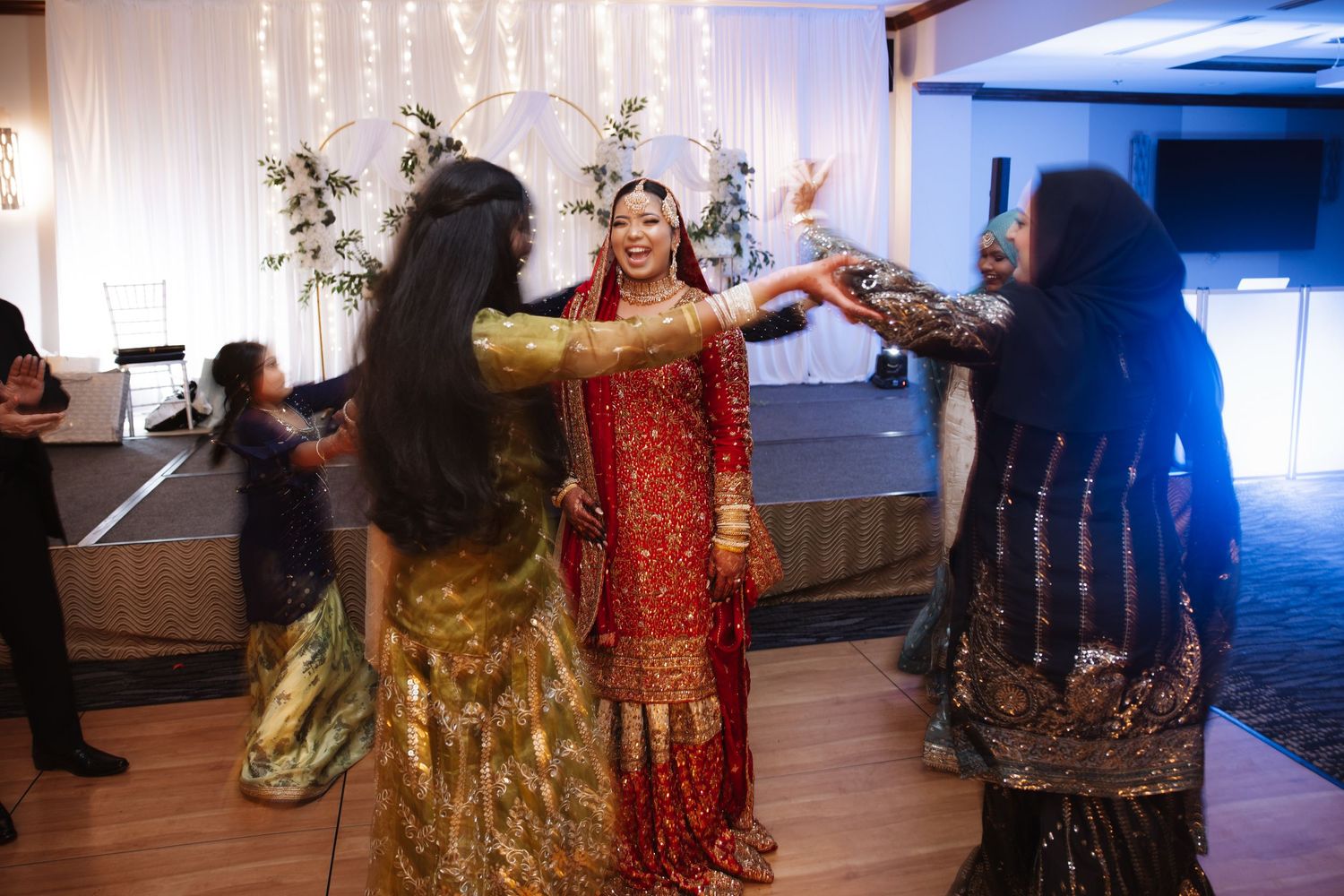 Bride in red lehenga dancing joyfully with guests in traditional Indian attire at wedding reception.