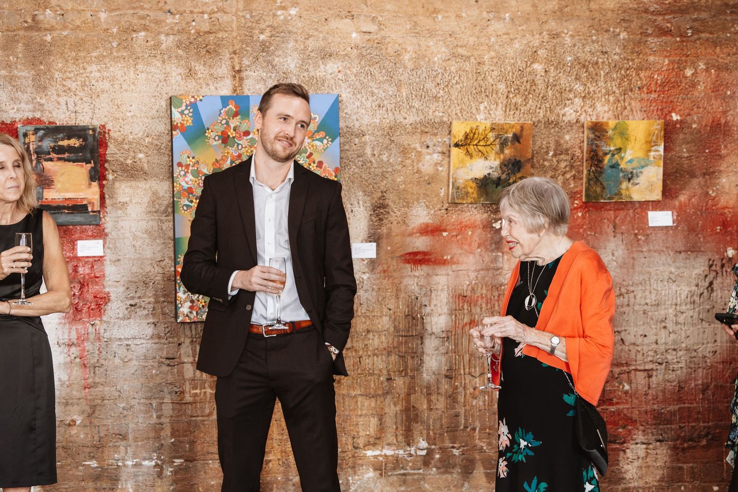Man and elderly woman conversing at an art gallery event with colorful paintings displayed on brick wall.