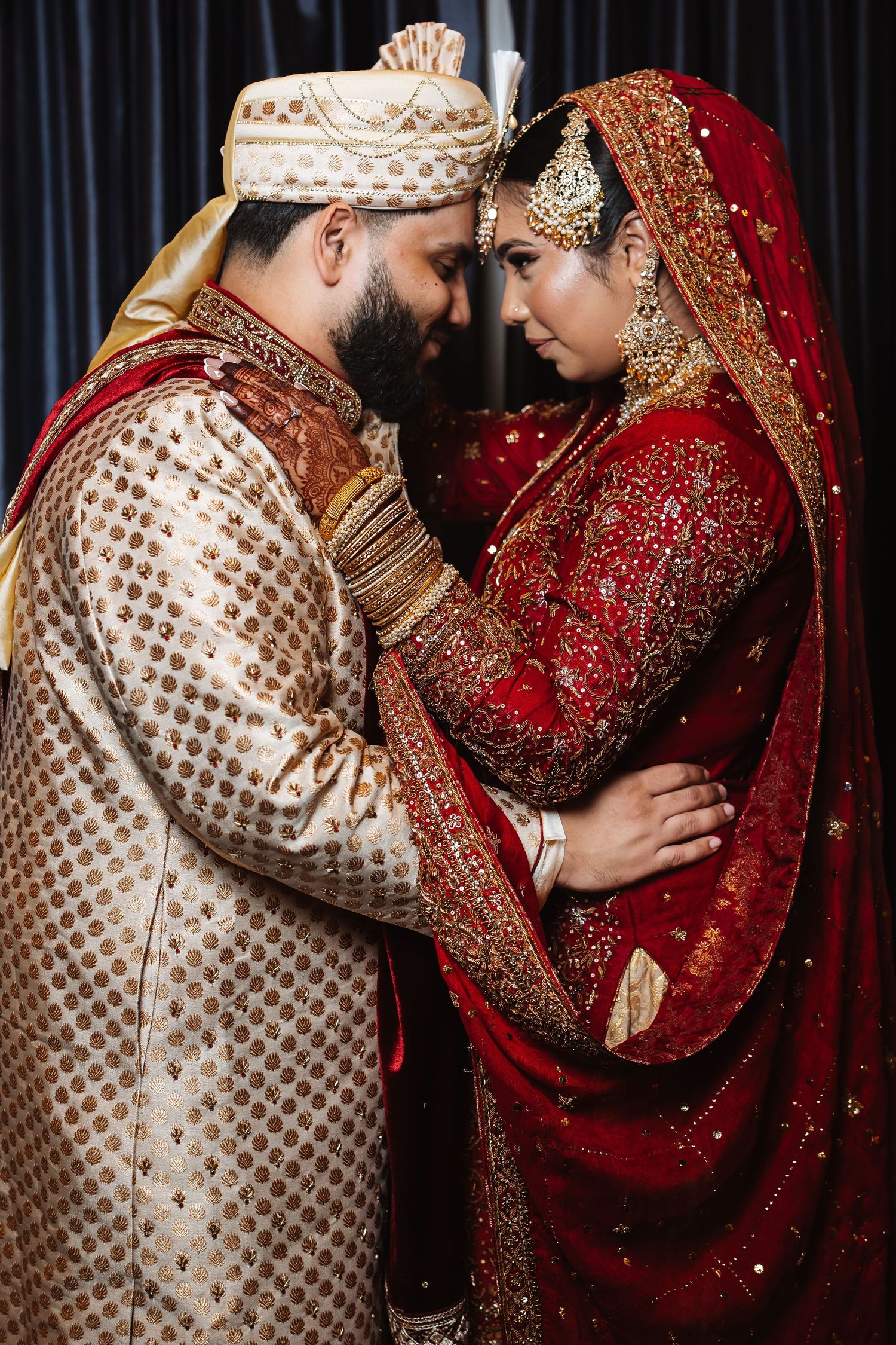 Indian bride and groom in traditional red and gold wedding attire sharing an intimate moment.
