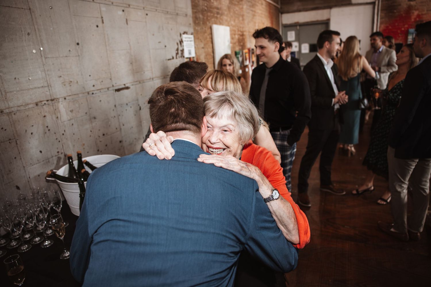 Smiling older woman in red hugs a guest in a blue suit at a festive event.