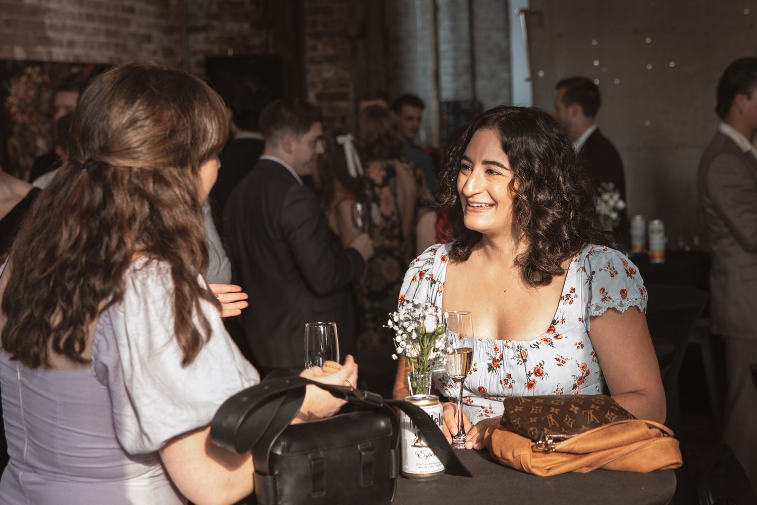 Two women in elegant dresses socializing and smiling at an indoor wedding reception event.