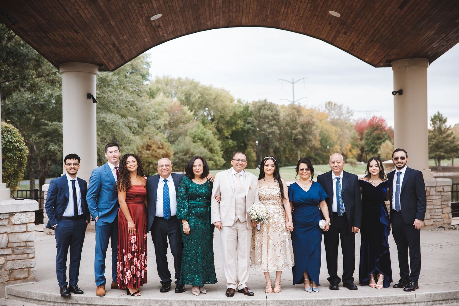 Wedding party group portrait under outdoor pavilion in formal attire with fall foliage.