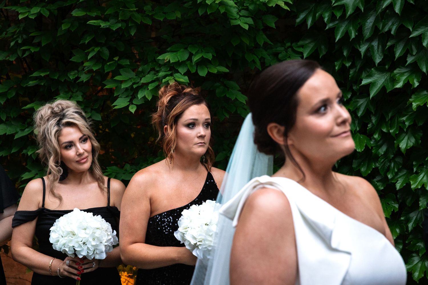 Bride in white gown with veil stands before bridesmaids in black dresses holding white bouquets at outdoor wedding.