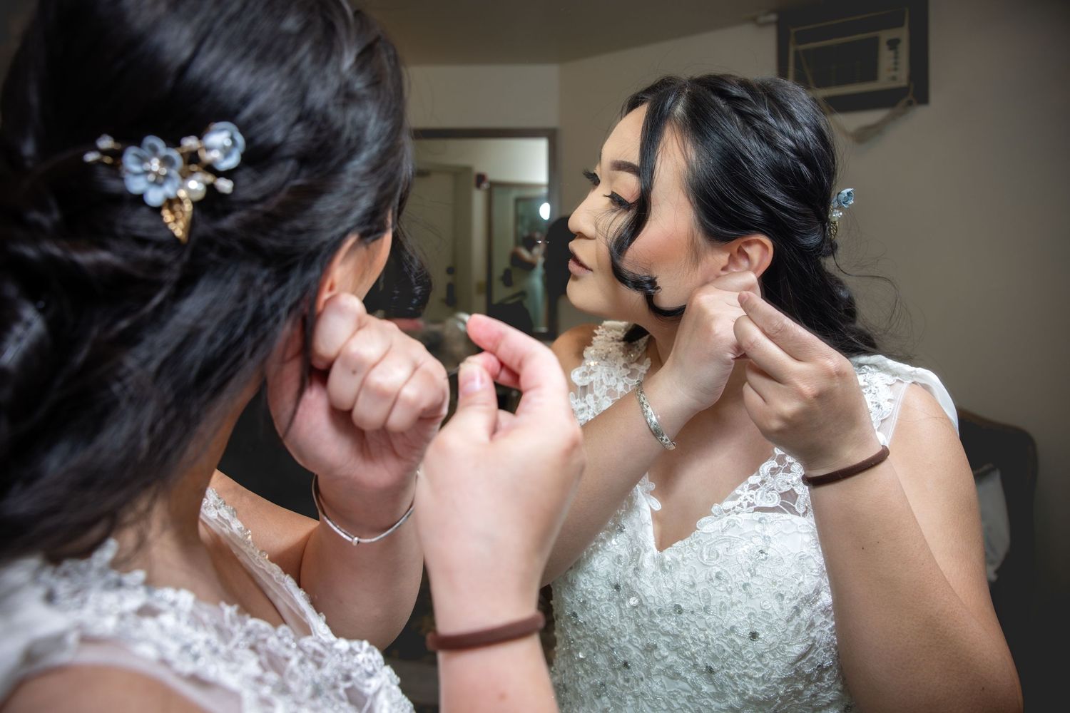 Bride getting earrings put on while wearing a lace wedding dress, reflected in a mirror.