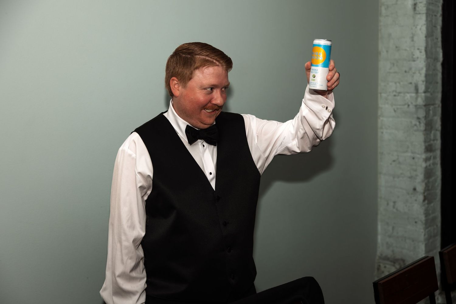 Smiling man in black vest and bow tie raises colorful can in celebratory toast at wedding.